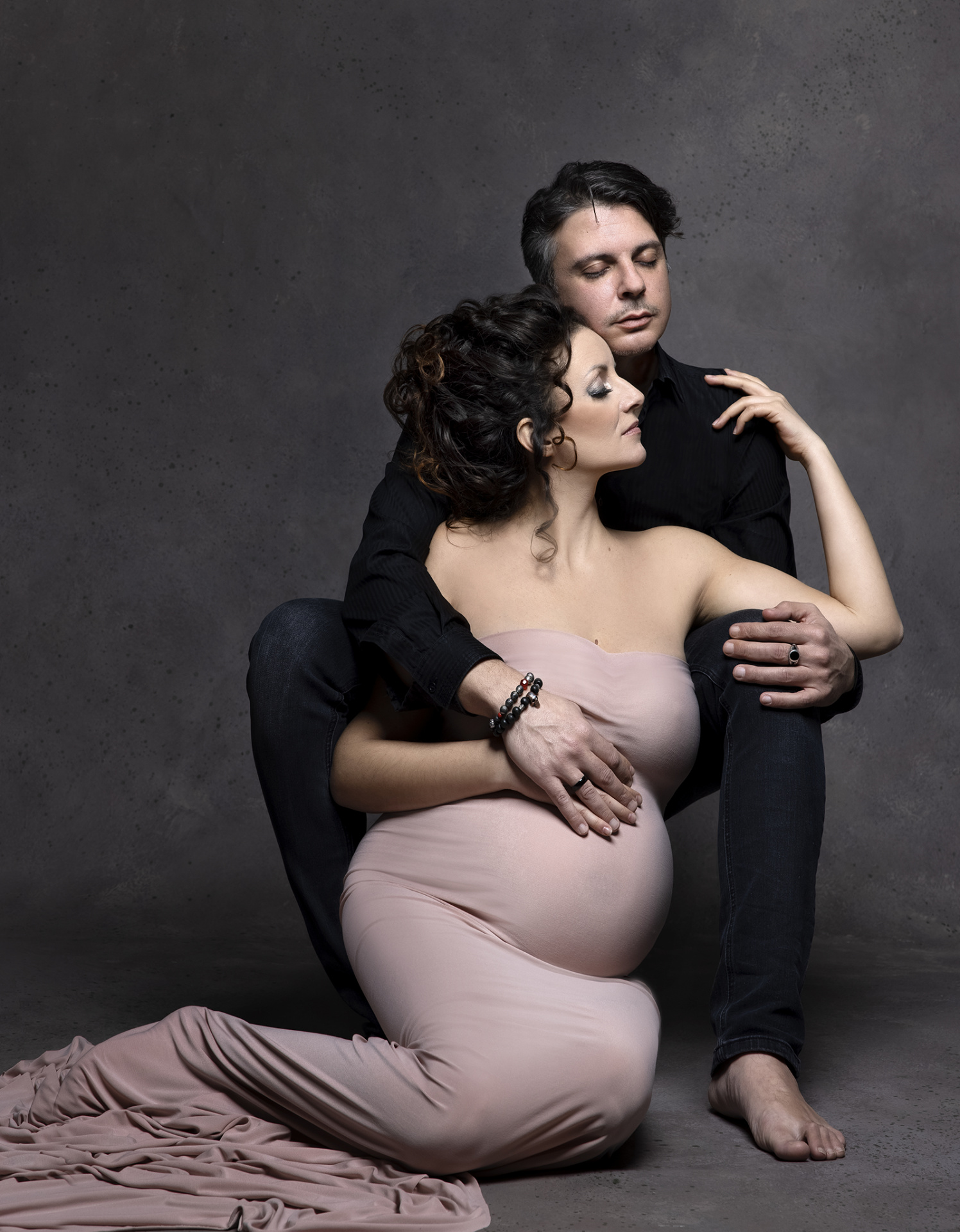 Servizio fotografico maternità a Bologna con Claudia Spaziani: coppia in posa intima, donna incinta in abito rosa e partner in nero, atmosfera elegante e affettuosa.