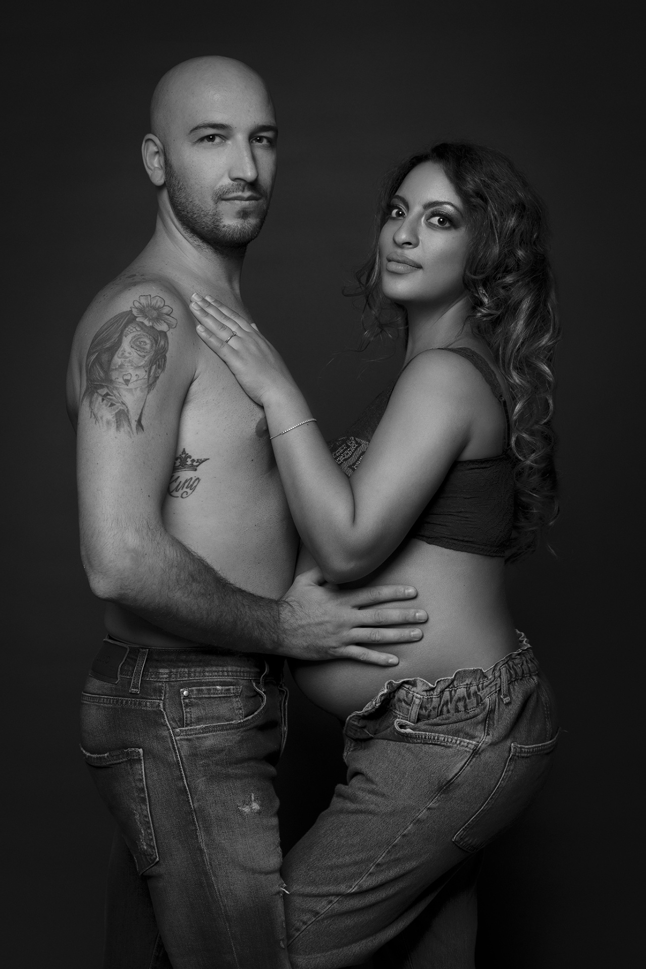 Ritratto di coppia maternità in bianco e nero a Bologna realizzato da Claudia Spaziani: donna incinta in jeans sbottonati e partner tatuato, posa affettuosa e intima.