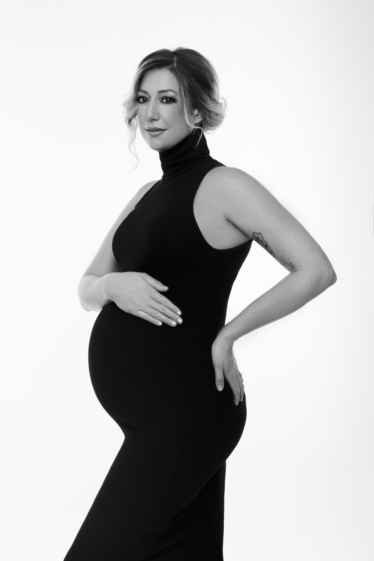 Foto maternità a Bologna firmata Claudia Spaziani: donna incinta in abito nero aderente, posa elegante e minimalista su sfondo bianco.