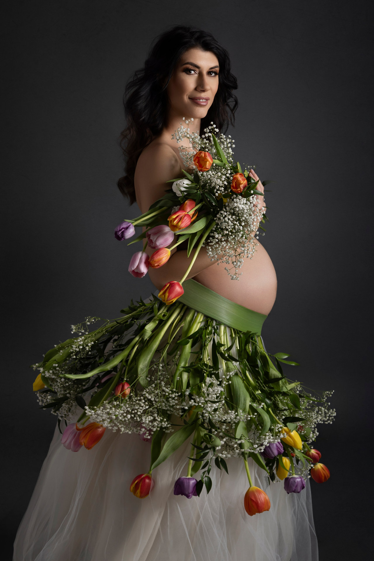 Foto maternità a Bologna firmata Claudia Spaziani: donna incinta adornata con tulipani e fiori variopinti, posa elegante e sorriso delicato.