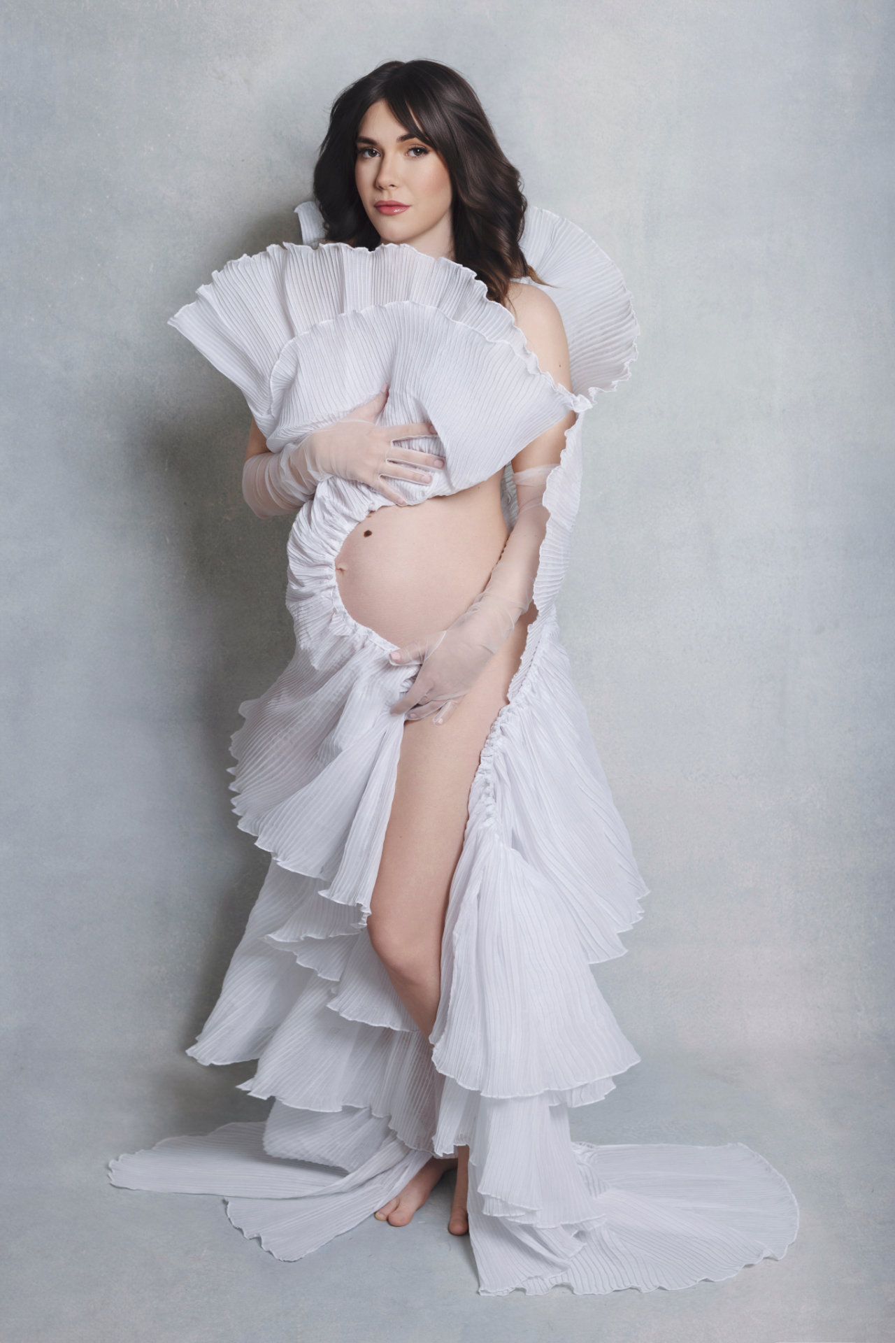 Foto maternità a Bologna firmata Claudia Spaziani: donna incinta in abito bianco plissettato con drappeggi voluminosi, posa artistica e luminosa.