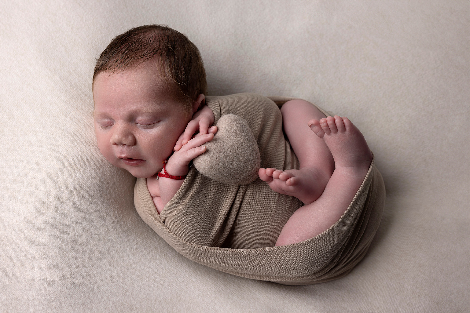 Ritratto di neonato maschio addormentato con cuscino a forma di cuore e braccialetto rosso, scattato nello studio fotografico di Claudia Spaziani a Bologna. Immagine tenera e simbolica.