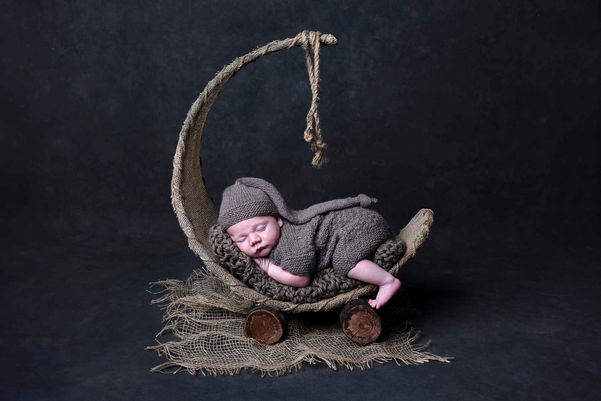 Ritratto creativo di neonato maschio addormentato in outfit grigio con cappellino, posato su culla a forma di luna, fotografato nello studio di Claudia Spaziani a Bologna. Immagine fiabesca e suggestiva.