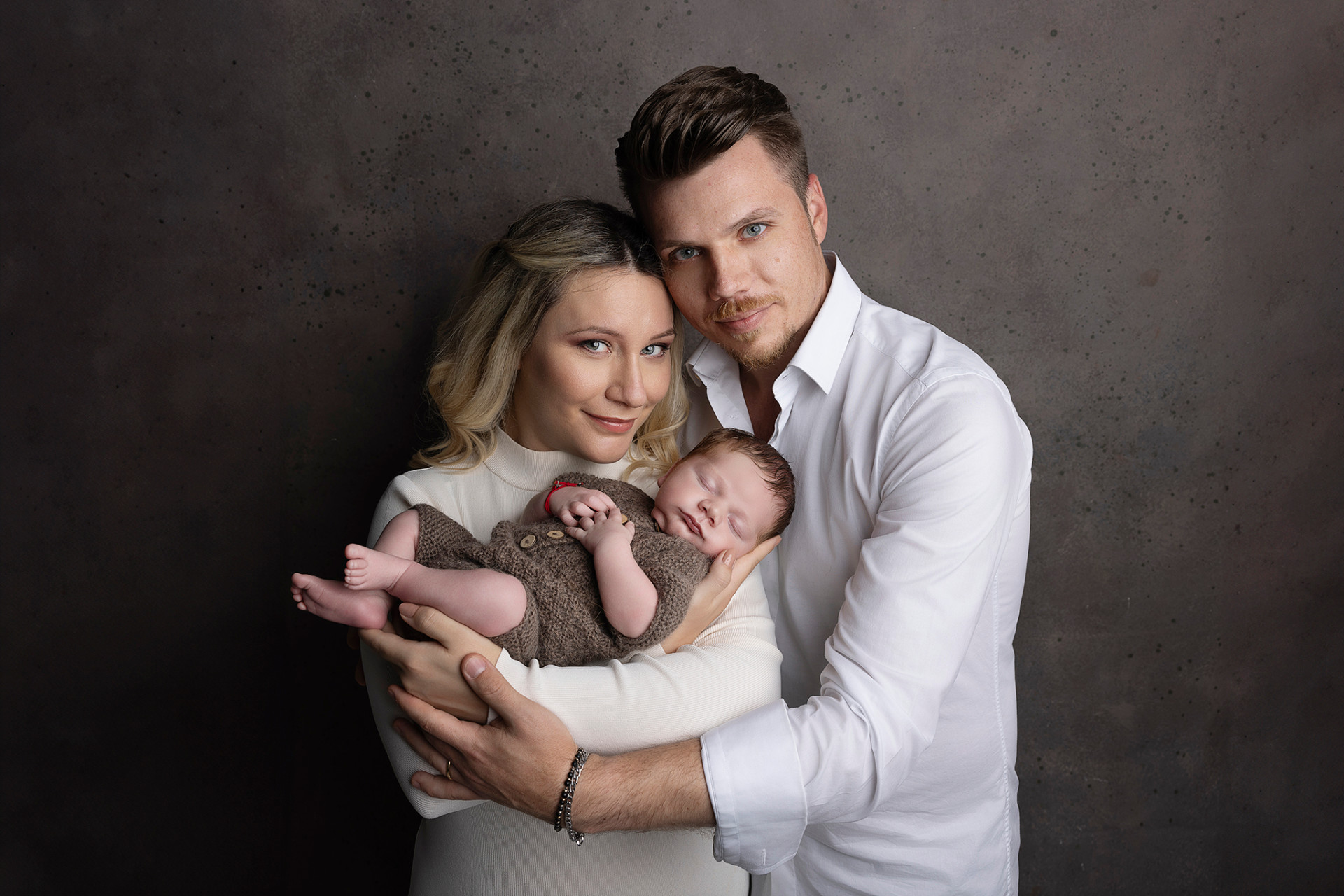 Foto di famiglia con neonato maschio addormentato in outfit marrone, scattata nello studio fotografico di Claudia Spaziani a Bologna. Immagine calda e affettuosa che celebra la nascita.