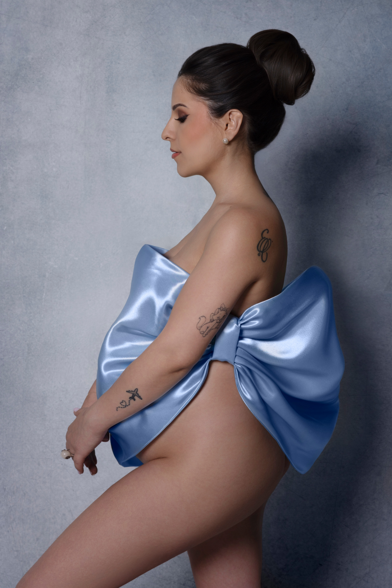 Donna incinta con grande fiocco di raso azzurro avvolto intorno al corpo, capelli raccolti e tatuaggi visibili. Fotografia artistica di Claudia Spaziani, Bologna.
