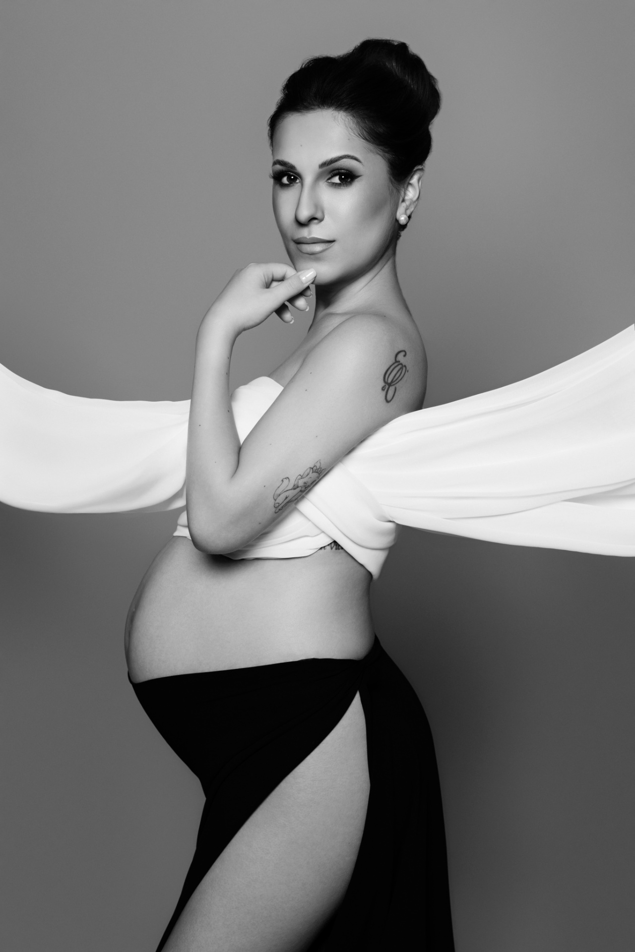 Donna incinta con tessuto bianco avvolto sul busto e gonna nera con spacco, posa raffinata con mano sul mento. Fotografia di Claudia Spaziani, Bologna.