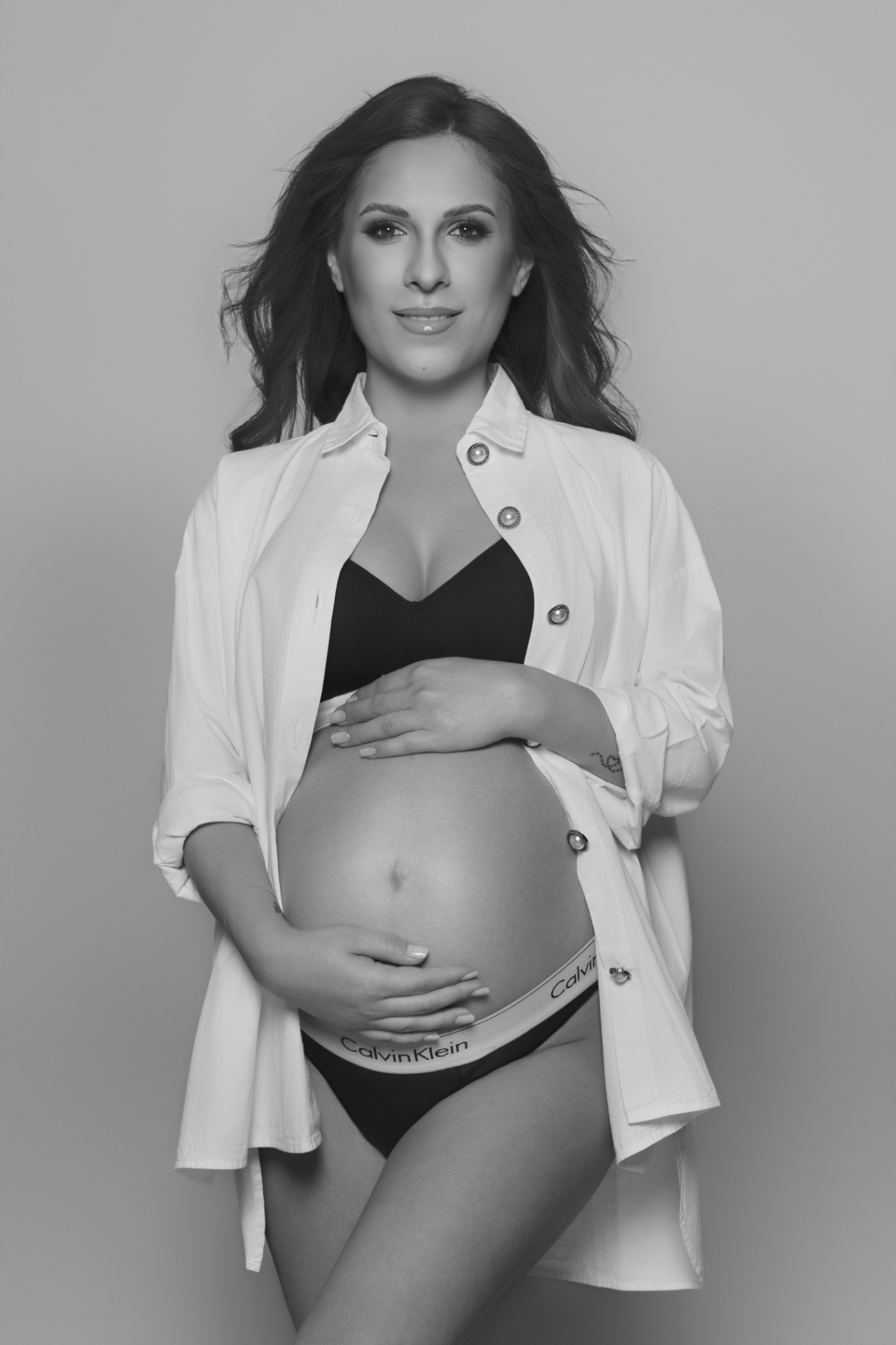 Donna incinta in reggiseno e intimo Calvin Klein con camicia bianca aperta, mani sulla pancia. Ritratto realizzato da Claudia Spaziani nel suo studio a Bologna.