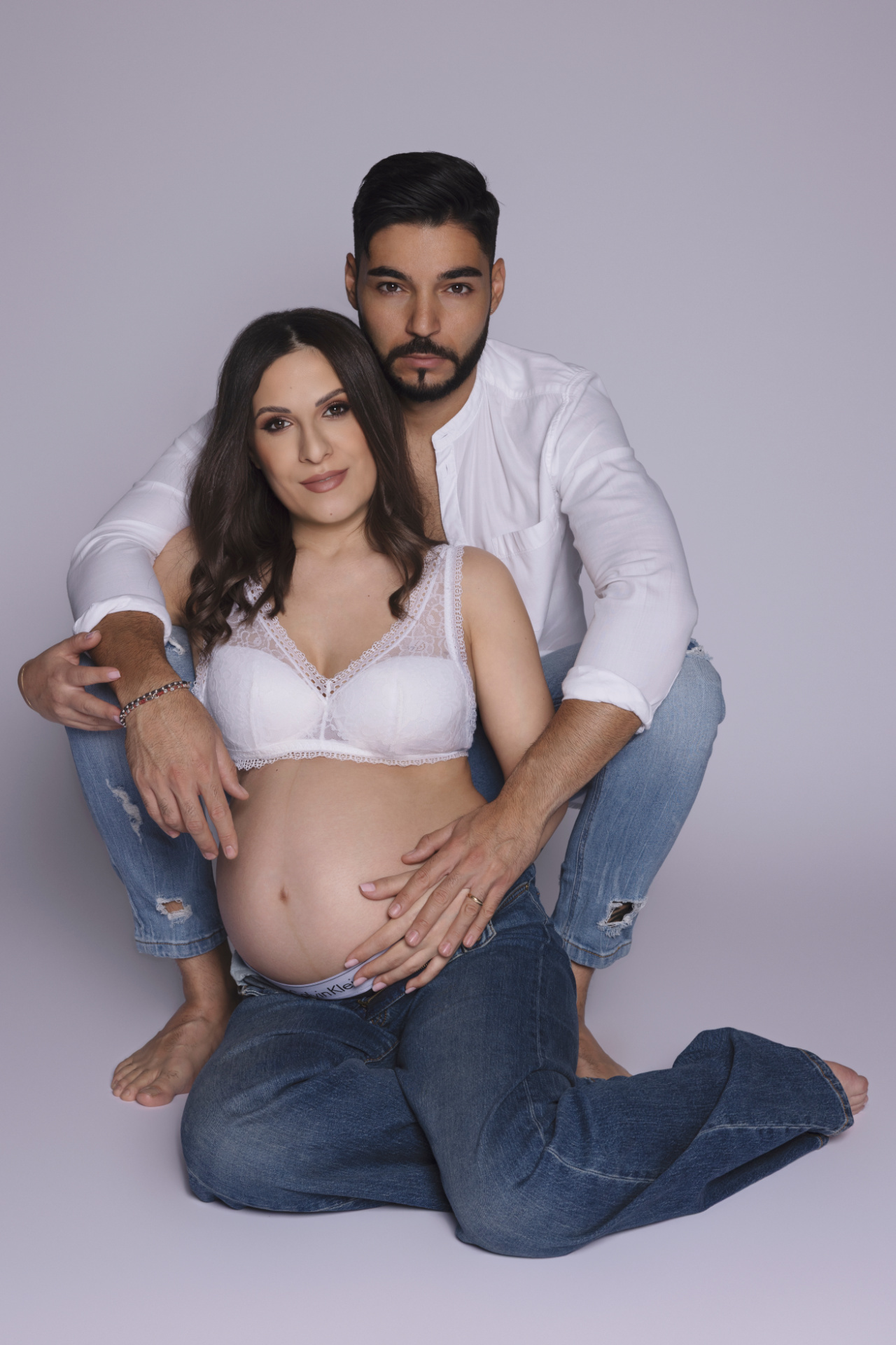 Donna incinta in bralette di pizzo bianco e jeans, abbracciata dal partner seduto dietro di lei, entrambi scalzi. Ritratto di maternità di Claudia Spaziani, Bologna.