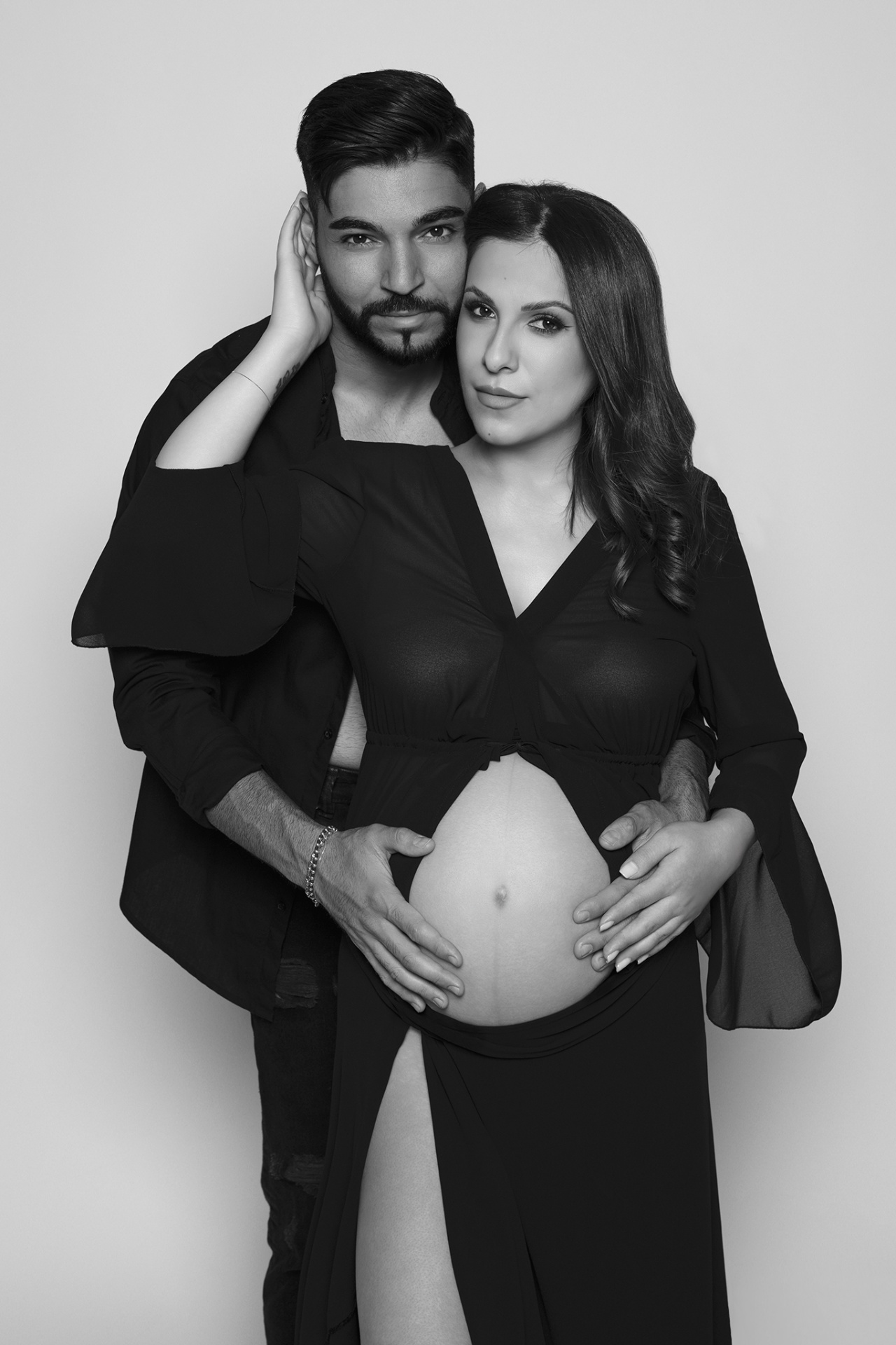 Donna incinta in abito lungo con spacco, partner dietro di lei con mani sulla pancia e gesto affettuoso sul volto. Foto di maternità di Claudia Spaziani, Bologna.