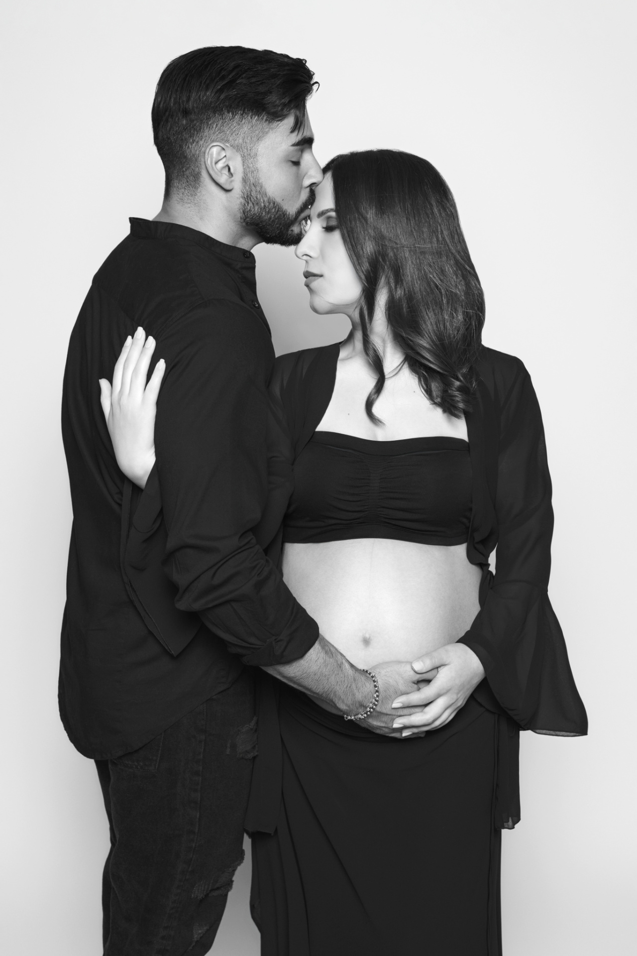 Donna incinta con top nero e gonna fluida, partner che le bacia la fronte e mani sulla pancia. Ritratto di Claudia Spaziani, Bologna.