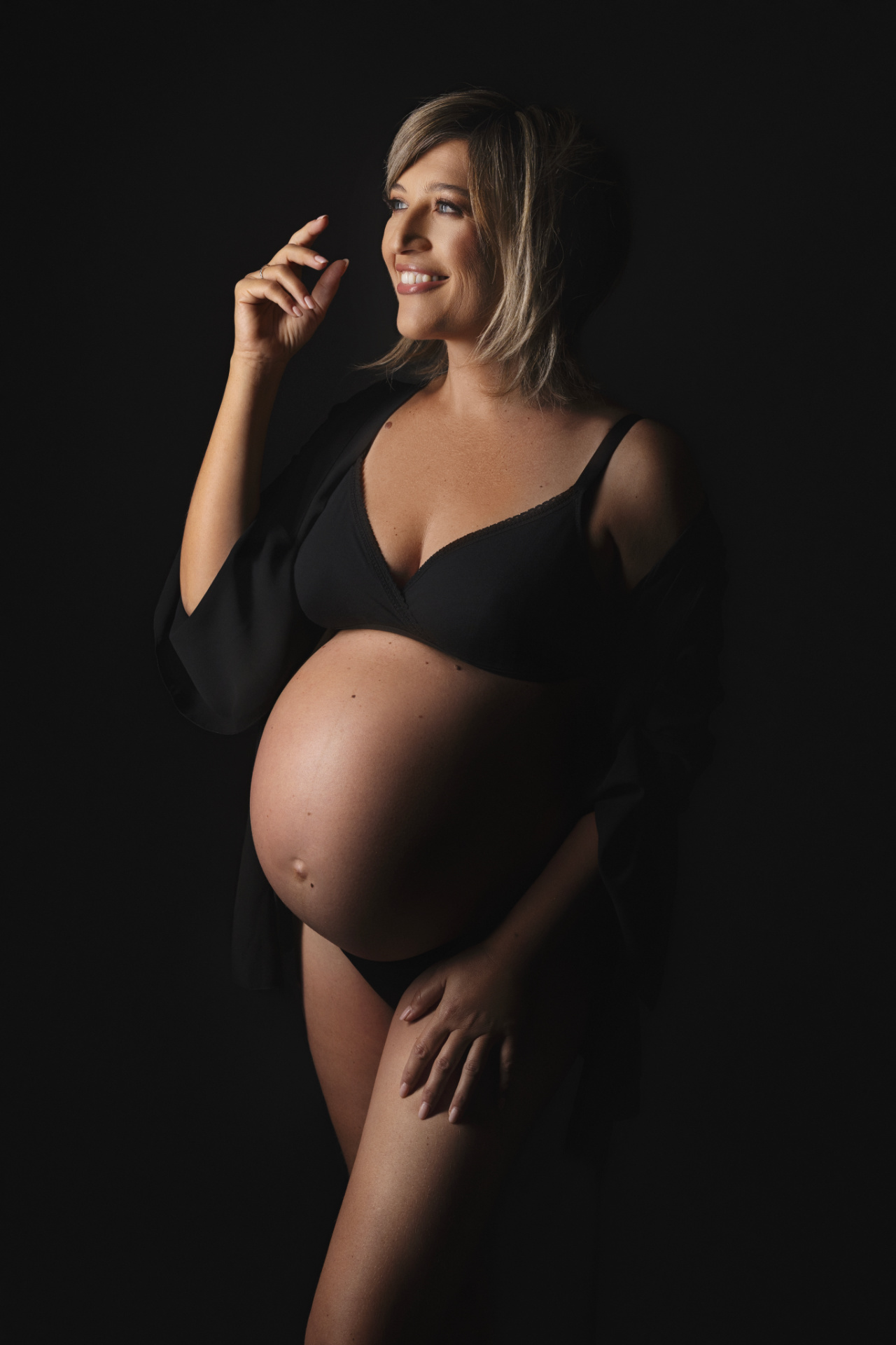 Donna incinta in lingerie nera con vestaglia, posa in piedi con mano sollevata verso il volto e sguardo rivolto in alto. Fotografia di Claudia Spaziani, Bologna.