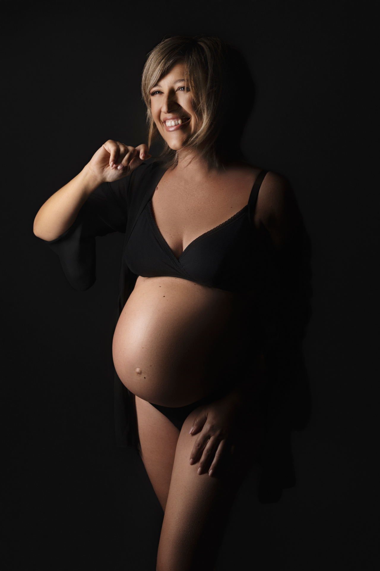 Donna incinta in reggiseno e intimo nero con vestaglia sulle spalle, illuminazione che mette in risalto il volto e la pancia. Ritratto di Claudia Spaziani, Bologna.