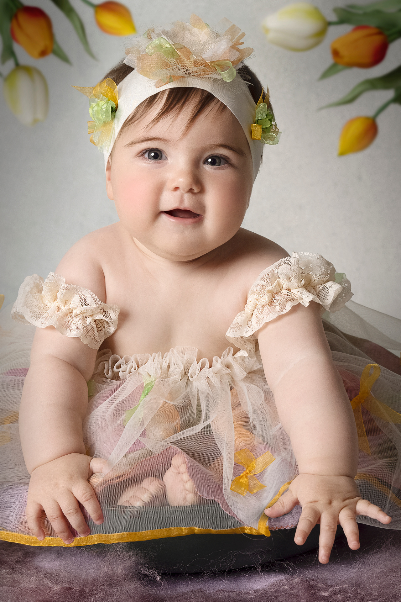 Ritratto delicato di bambina di 6 mesi in abito con fiocchi gialli e fascia floreale, circondata da tulipani, realizzato da Claudia Spaziani a Bologna.