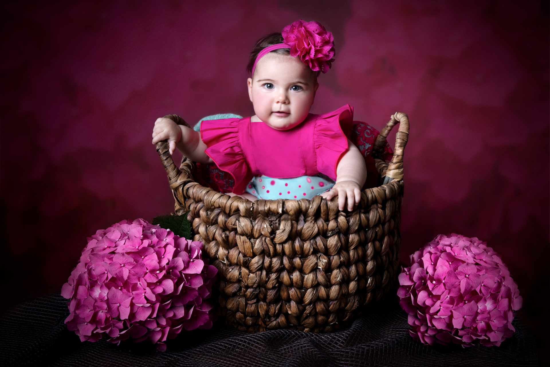 Ritratto professionale di bambina di 6 mesi in abito rosa con fascia floreale, seduta in cesto intrecciato, scattato da Claudia Spaziani nel suo studio fotografico a Bologna.