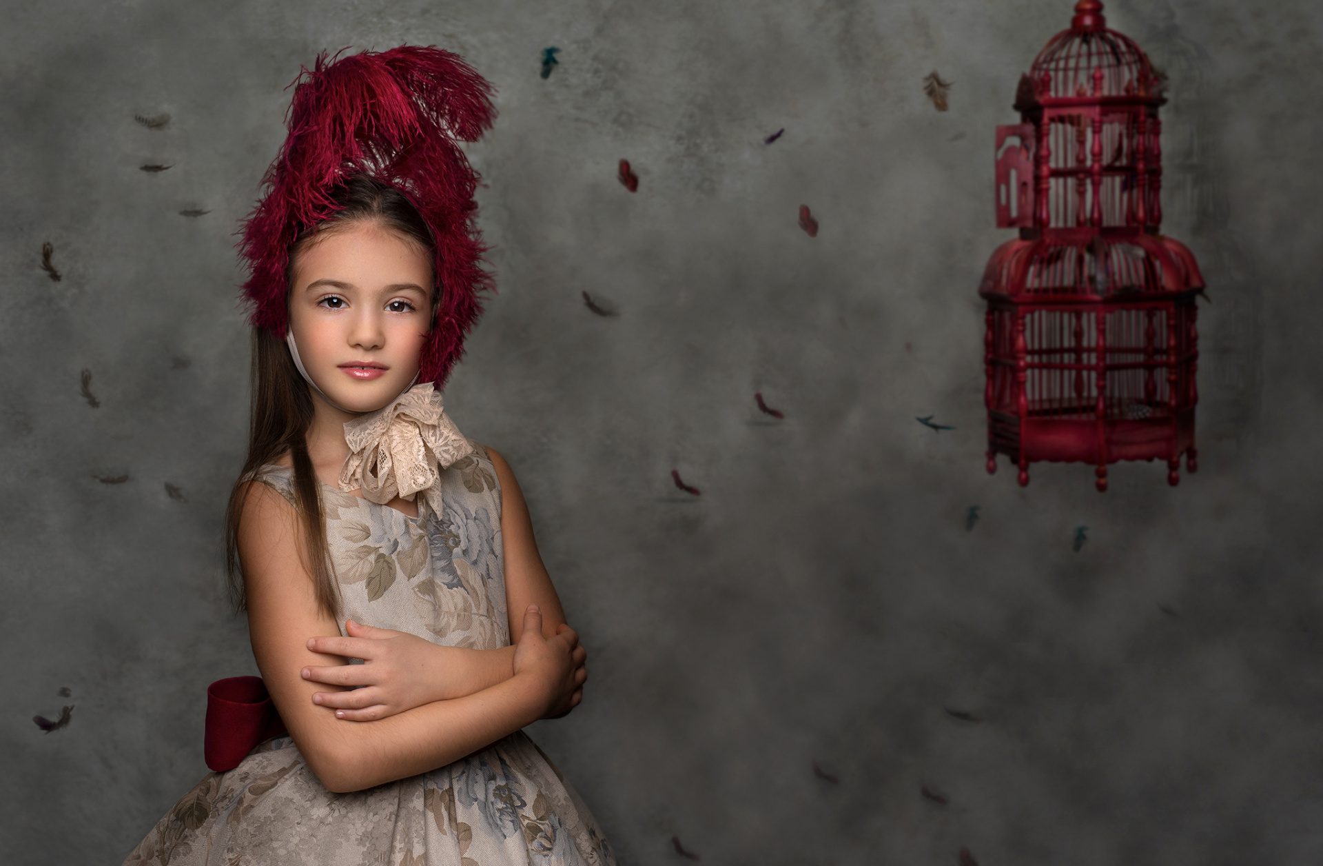 Ritratto concettuale di bambina in abito vintage floreale con copricapo di piume rosse e gabbietta decorativa, scattato da Claudia Spaziani nel suo studio fotografico a Bologna.