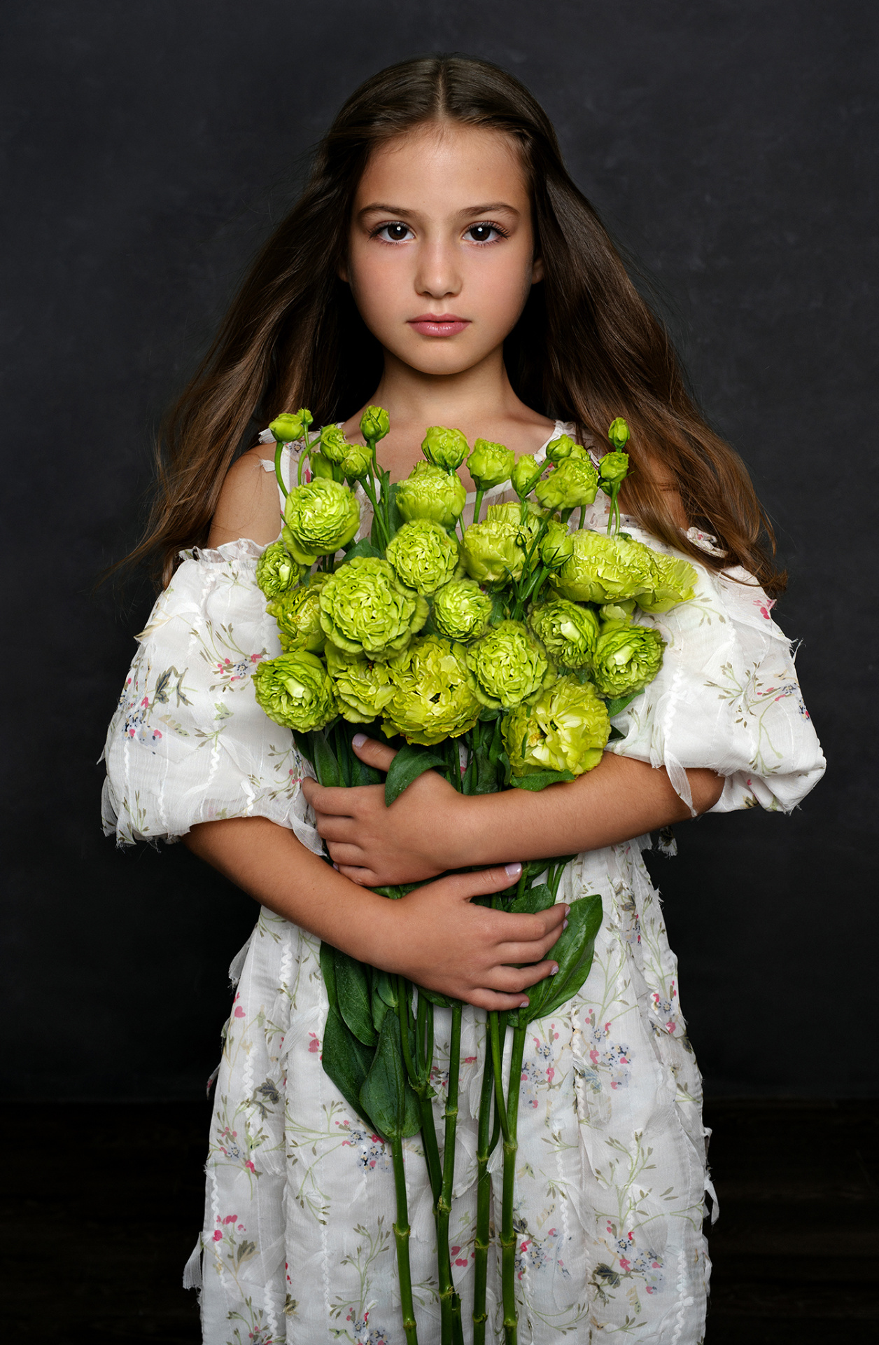 Ritratto elegante di bambina con abito floreale e bouquet di ranuncoli verdi, su sfondo scuro, realizzato da Claudia Spaziani nel suo studio fotografico a Bologna.