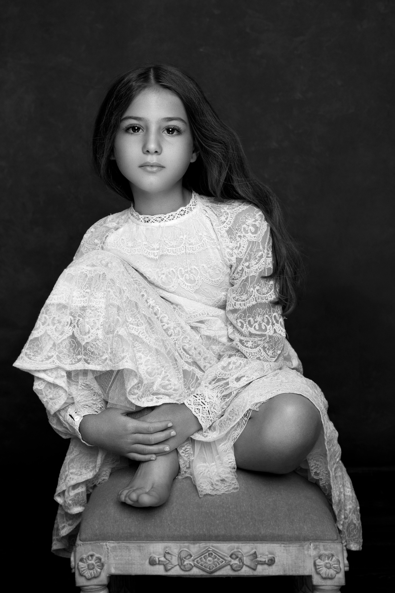 Ritratto fine-art in bianco e nero di bambina in abito lungo di pizzo, seduta su sgabello decorato, scattato da Claudia Spaziani nel suo studio fotografico a Bologna.