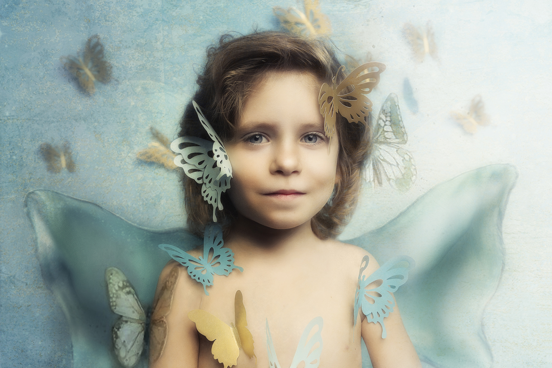 Ritratto etereo di bambino con ali trasparenti e farfalle colorate, su sfondo blu, scattato da Claudia Spaziani nel suo studio fotografico a Bologna.