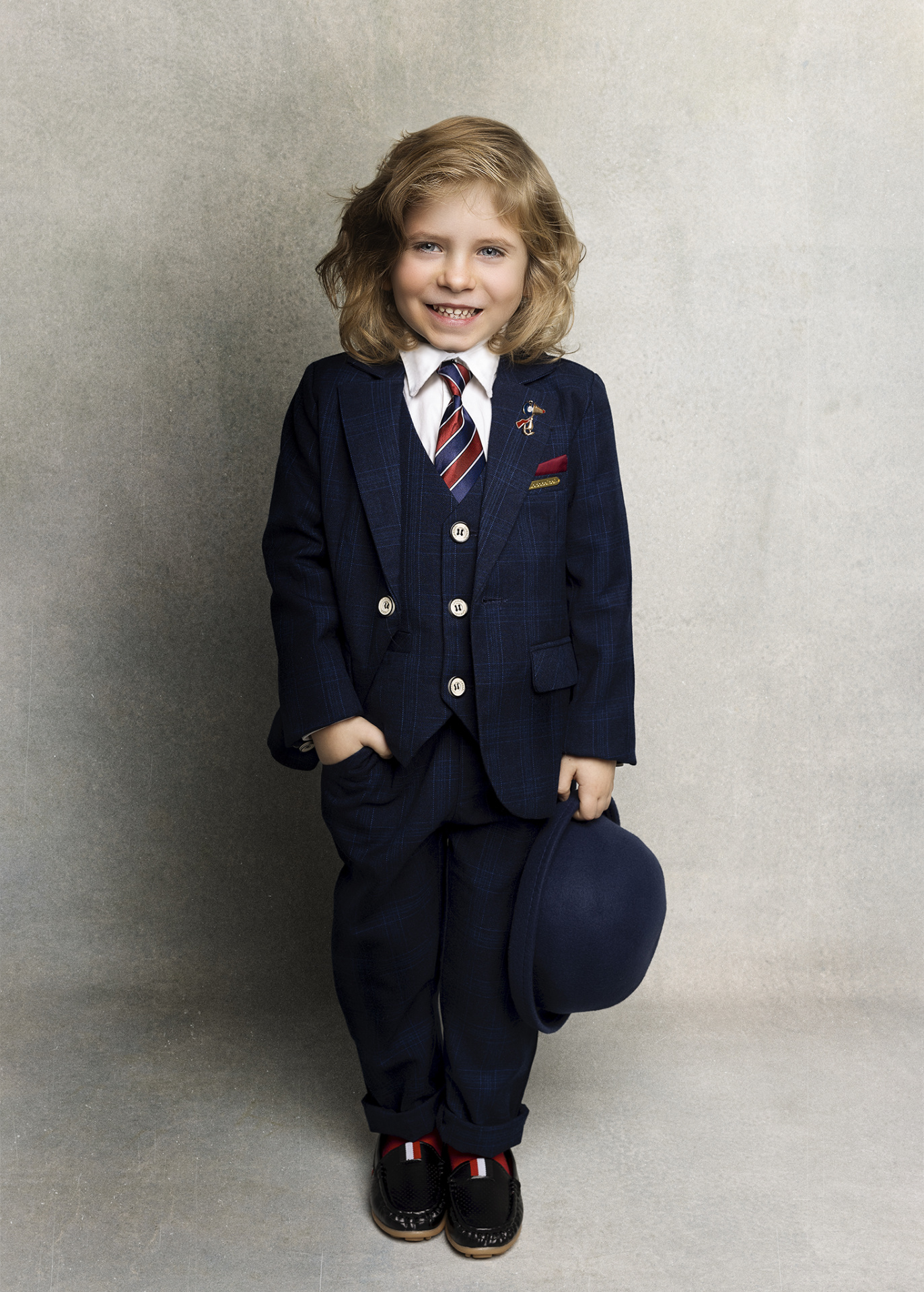 Ritratto formale di bambino in abito blu con cravatta rossa, bianca e blu, e bombetta, realizzato da Claudia Spaziani nel suo studio fotografico a Bologna.