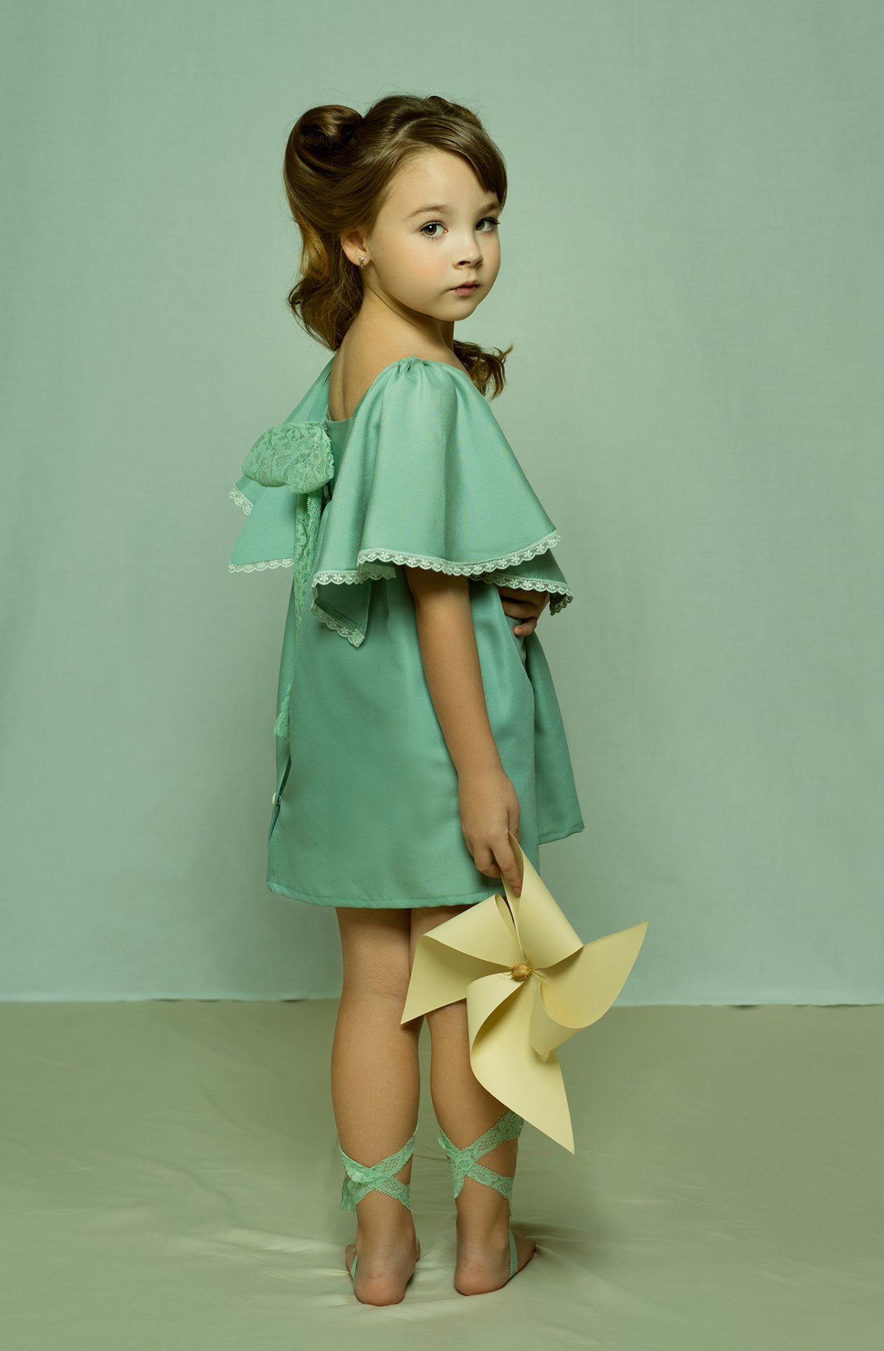 Ritratto delicato di bambina in abito verde menta con fiocco e girandola, su sfondo pastello, scattato da Claudia Spaziani nel suo studio fotografico a Bologna.