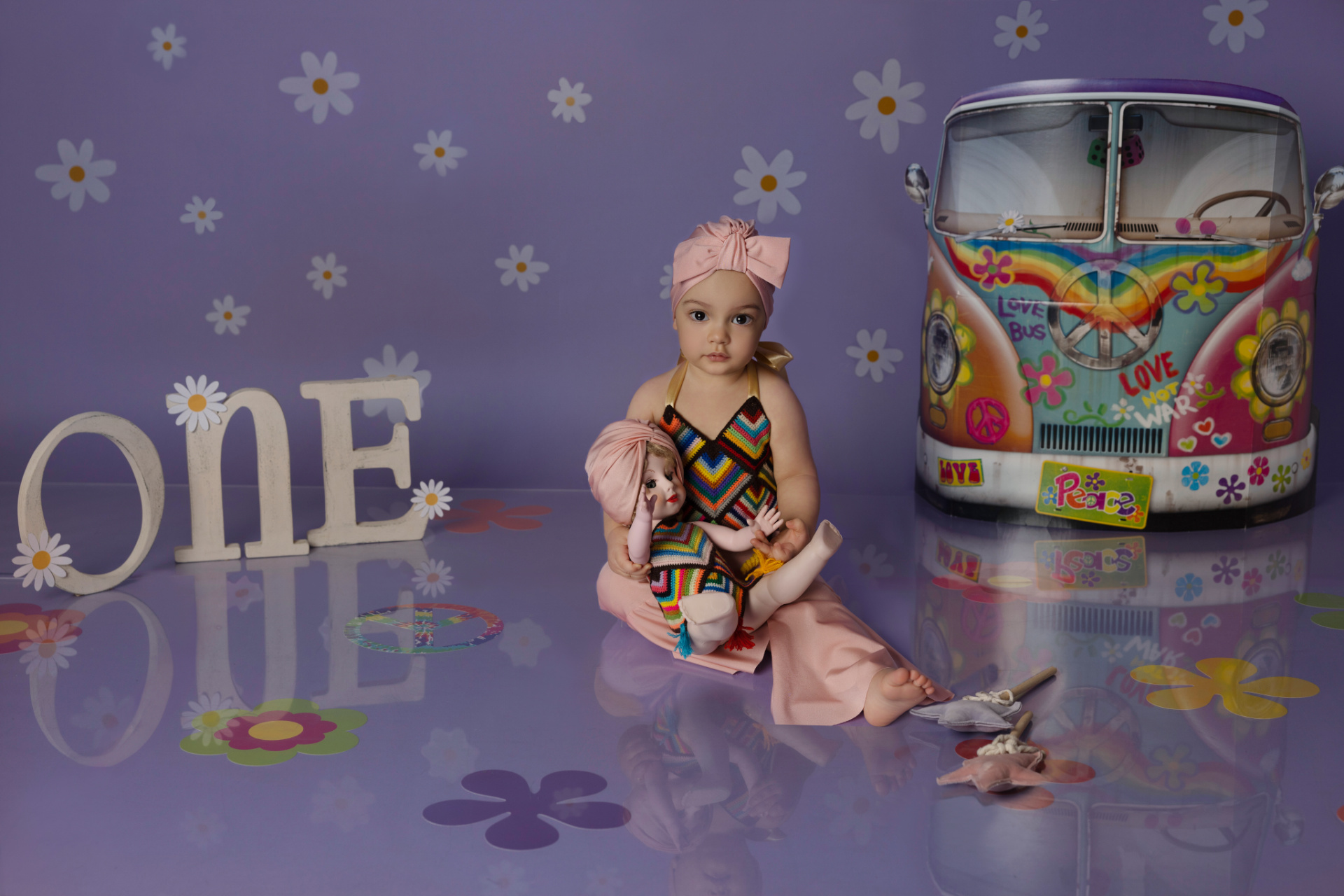 Bambina seduta su pavimento viola decorato con fiori, accanto a una bambola vestita in stile hippie. Lettere "ONE" con margherite e furgoncino colorato con scritte "LOVE BUS" e "Peace". Ritratto di primo compleanno realizzato da Claudia Spaziani nel suo studio fotografico a Bologna.
