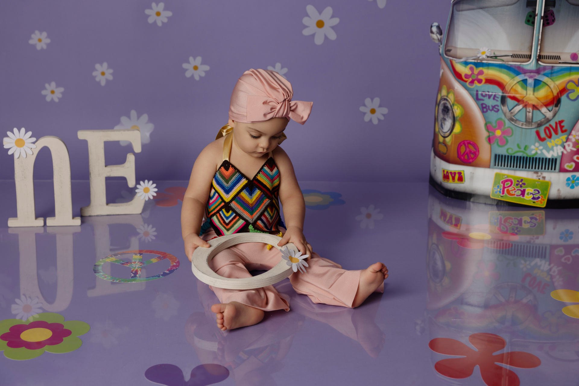 Bambina con fascia rosa e abbigliamento a zigzag colorato, seduta su pavimento viola con fiori e lettere "ONE" decorate. Sfondo con furgone hippie e scritte "LOVE NOT WAR". Foto smash cake di Claudia Spaziani, fotografa ritrattista a Bologna.