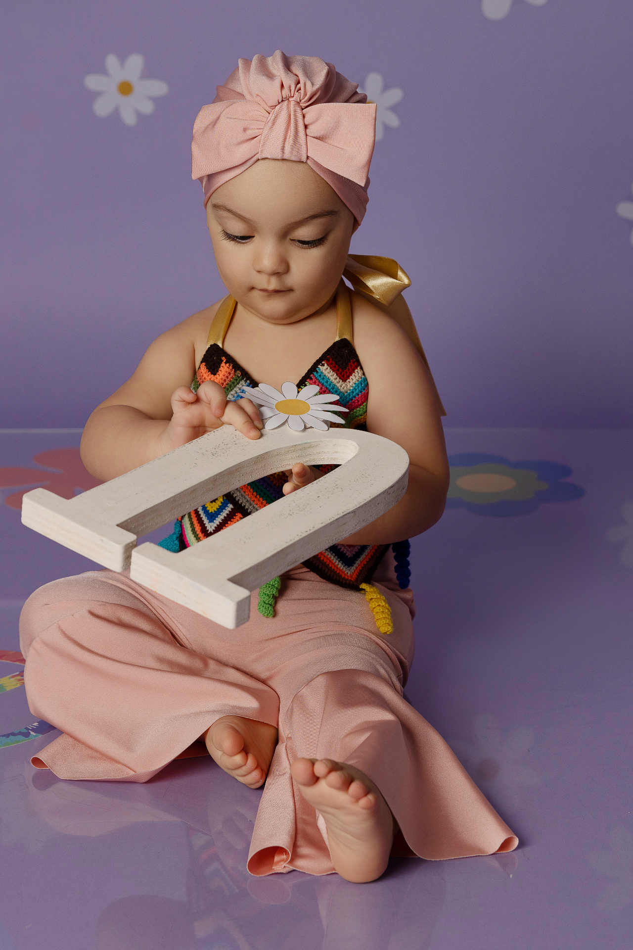 Bambina con fascia rosa e top all’uncinetto colorato, seduta su pavimento viola con fiori, mentre tiene la lettera "U" decorata con margherita. Ritratto creativo di primo compleanno realizzato da Claudia Spaziani nel suo studio fotografico a Bologna.