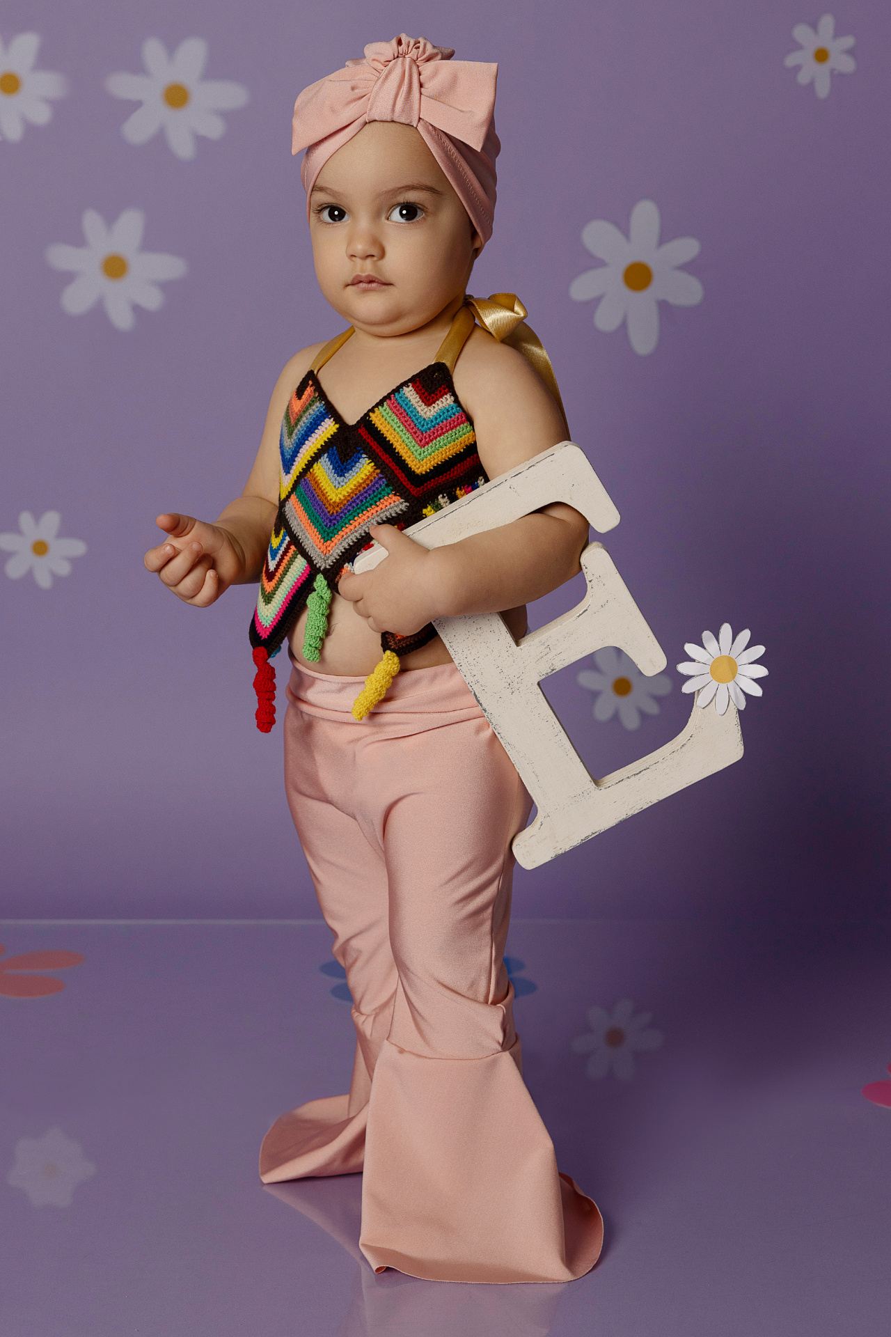 Bambina in abbigliamento boho con pantaloni rosa e top a zigzag, in piedi davanti a sfondo viola con margherite, mentre tiene la lettera "E". Foto di primo compleanno in stile hippie scattata da Claudia Spaziani a Bologna.