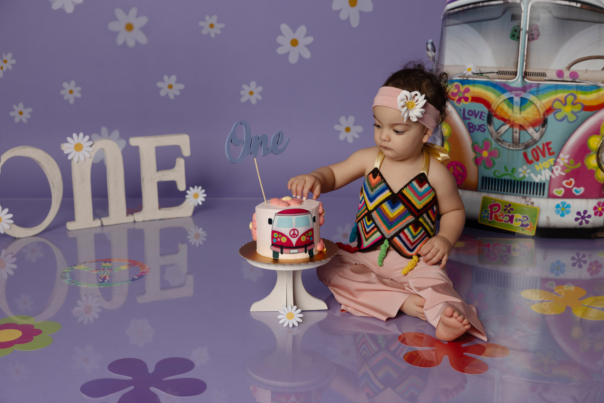Bambina con fascia rosa e vestito colorato seduta davanti a torta decorata con furgone "Peace" e topper "One". Lettere "ONE" e sfondo hippie con scritte "LOVE BUS". Ritratto smash cake di Claudia Spaziani, fotografa professionista a Bologna.