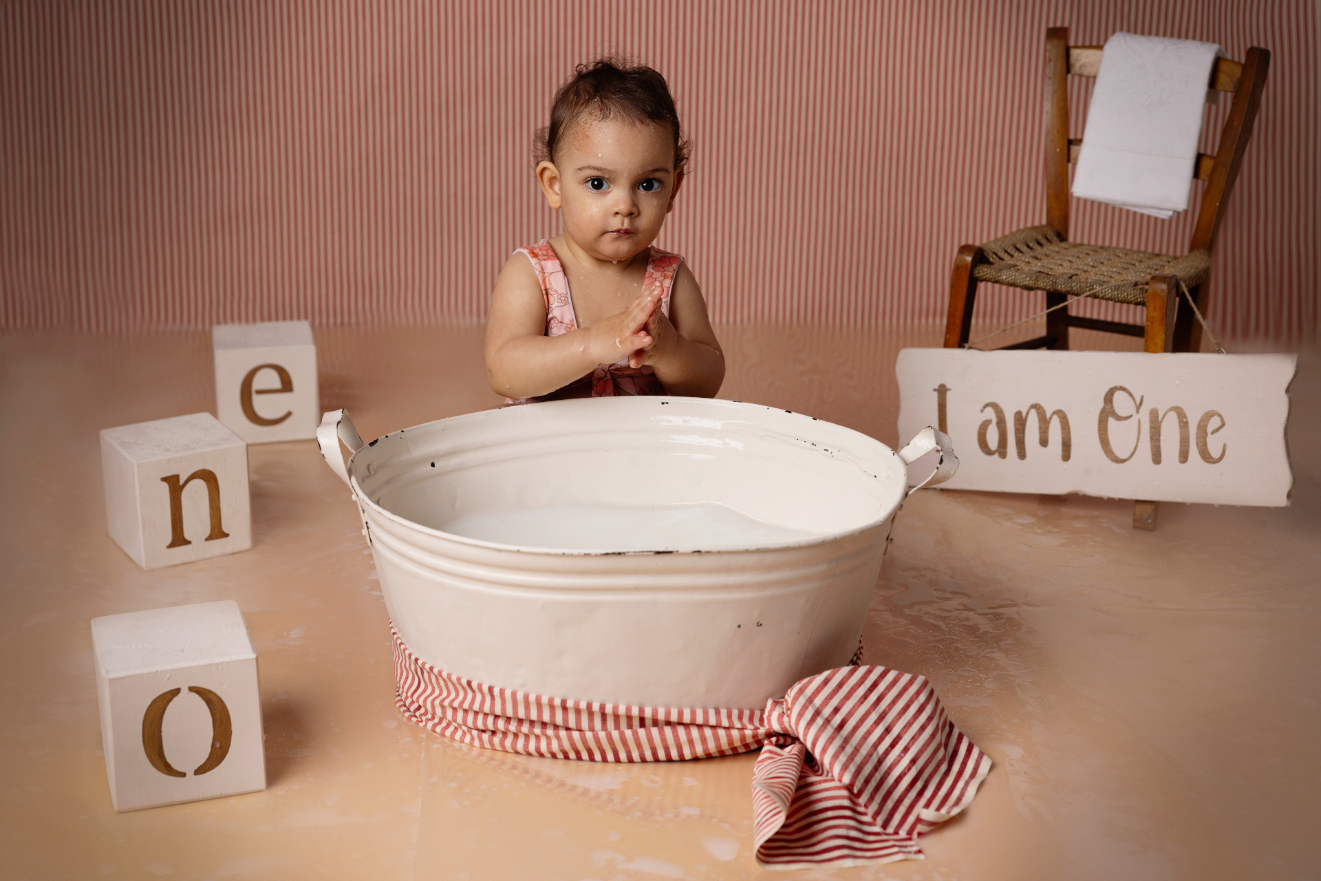 Bambina in vasca bianca per milk bath, con outfit rosa e blocchi in legno che formano "One". Sfondo a righe rosse e bianche, con cartello "I am One". Foto di primo compleanno creativa scattata da Claudia Spaziani nel suo studio fotografico a Bologna.