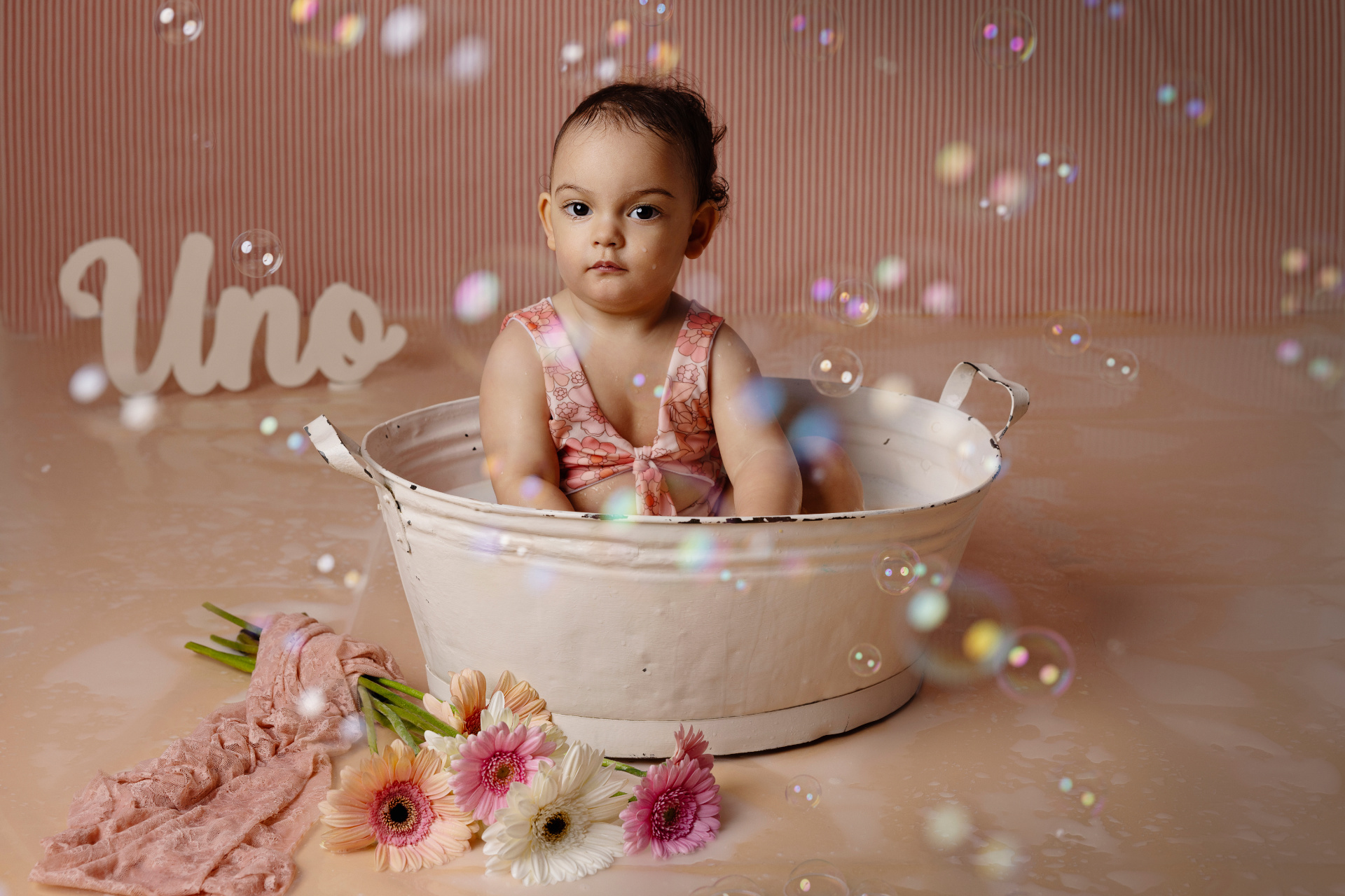 Bambina in vasca bianca con bolle di sapone, outfit floreale rosa e sfondo a righe rosa con scritta "Uno". Fiori gerbera colorati in primo piano. Ritratto di primo compleanno realizzato da Claudia Spaziani a Bologna.