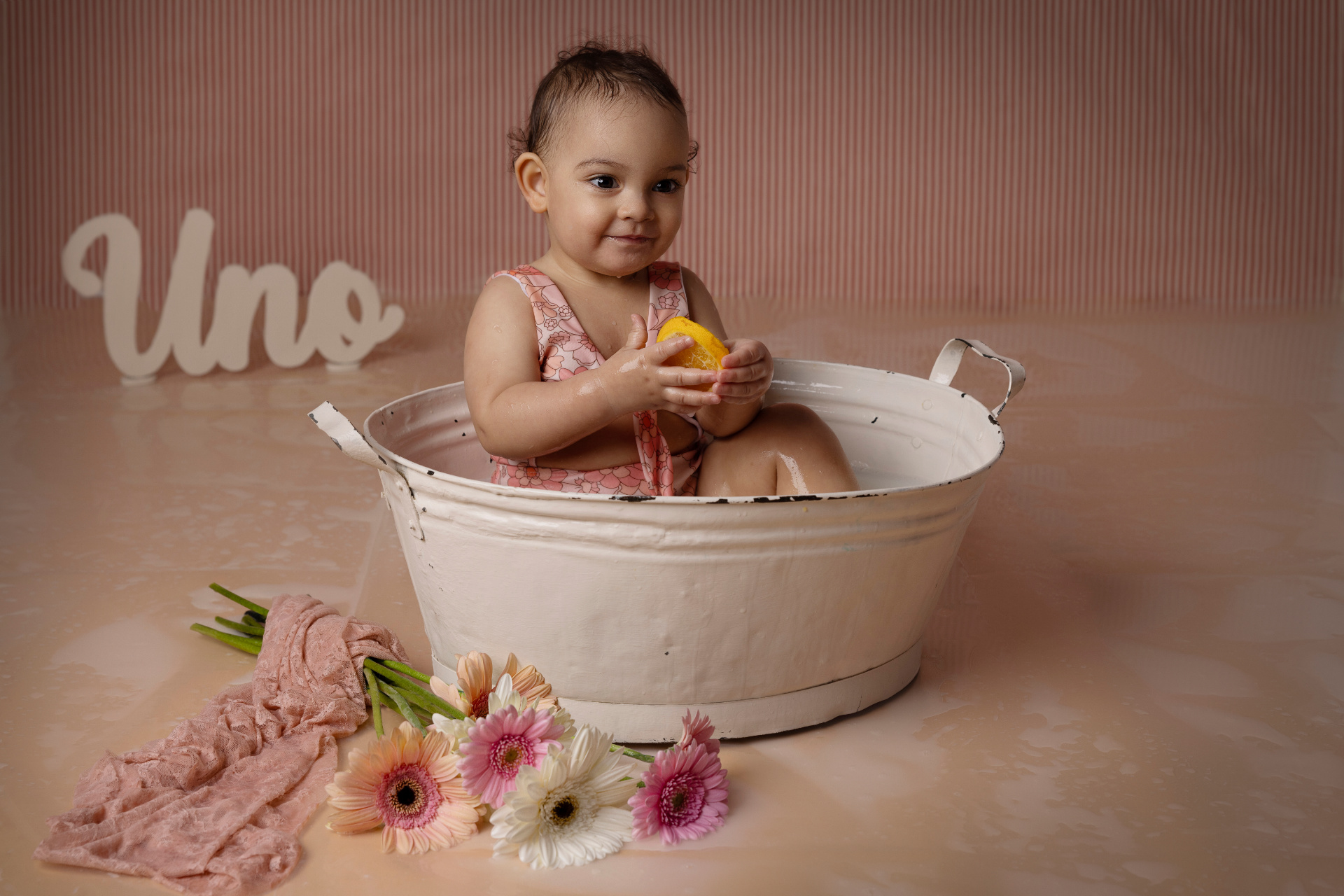Bambina in vasca bianca con fetta d’arancia, vestita in rosa floreale, davanti a scritta "Uno" e fiori gerbera. Foto di primo compleanno in stile milk bath scattata da Claudia Spaziani nel suo studio a Bologna.