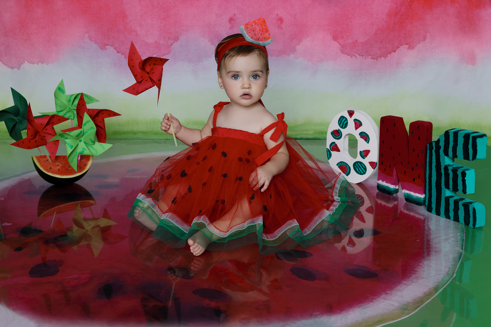 Bambina con vestito a tema anguria e girandola rossa, seduta davanti a lettere "ONE" decorate. Sfondo rosa, bianco e verde con motivi di anguria. Ritratto di primo compleanno realizzato da Claudia Spaziani nel suo studio fotografico a Bologna.