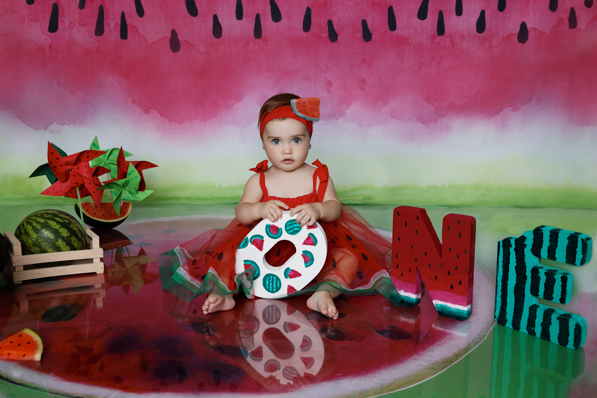 Bambina con vestito rosso e fascia a tema anguria, seduta su pavimento decorato con angurie e lettere "ONE". Sfondo dipinto con motivi di anguria. Ritratto di smash cake realizzato da Claudia Spaziani nel suo studio fotografico a Bologna.