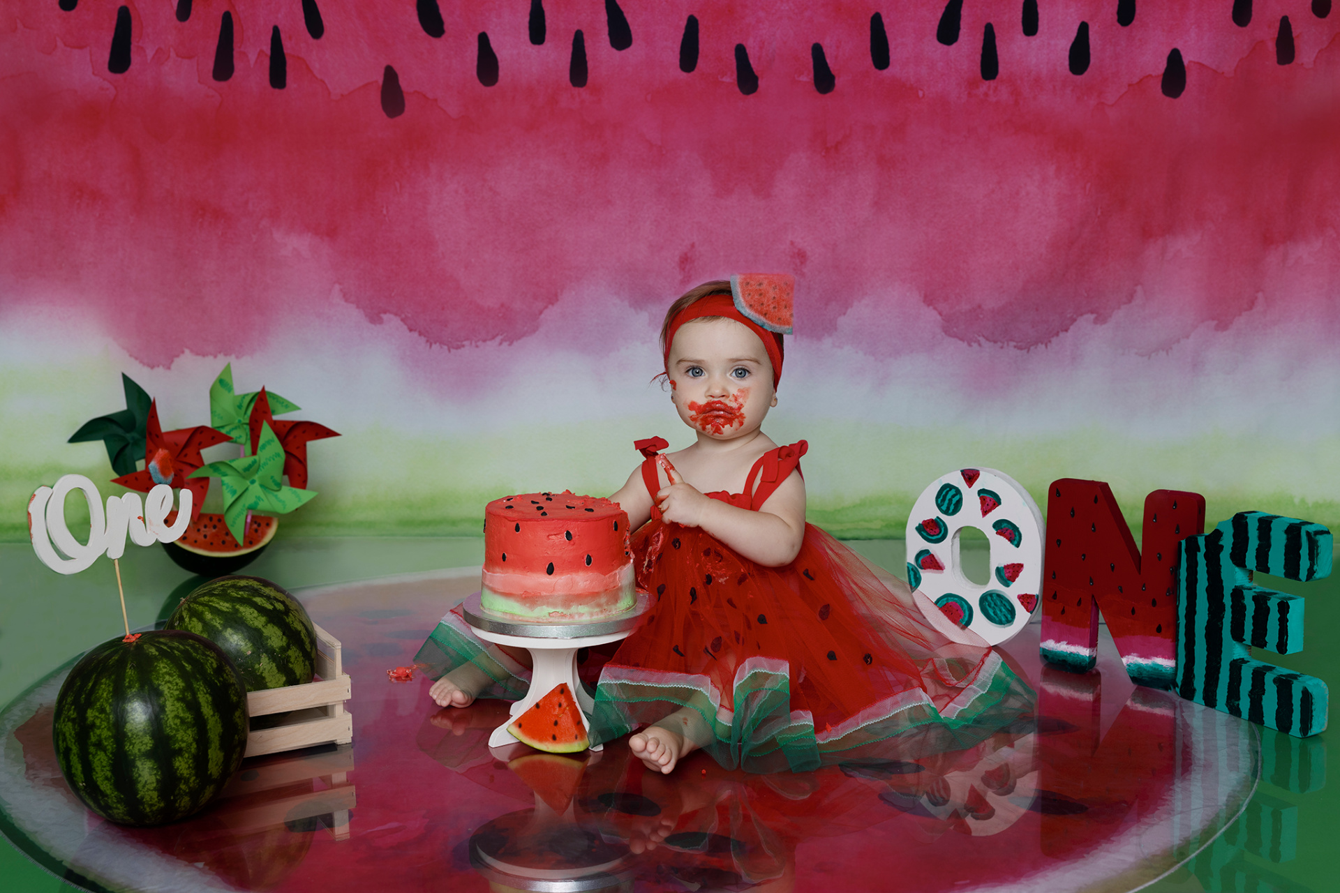 Bambina con vestito a tema anguria e torta parzialmente mangiata, seduta su superficie riflettente con sfondo anguria. Lettere "ONE" e decorazioni festive. Ritratto smash cake di Claudia Spaziani, fotografa professionista a Bologna.