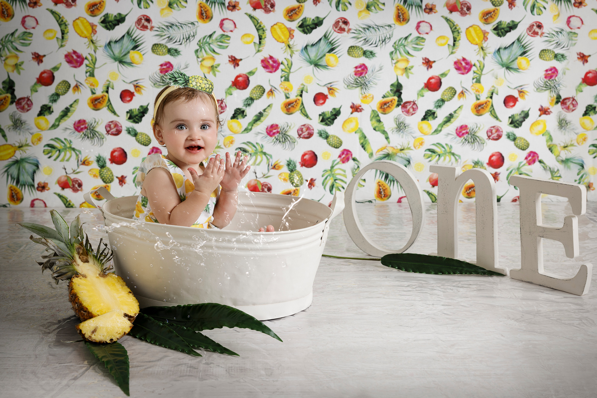 Bambina seduta in vasca bianca con sfondo tropicale decorato con frutti colorati. Lettere "ONE" e bolle di sapone completano la scena. Ritratto di primo compleanno tropicale scattato da Claudia Spaziani nel suo studio fotografico a Bologna.