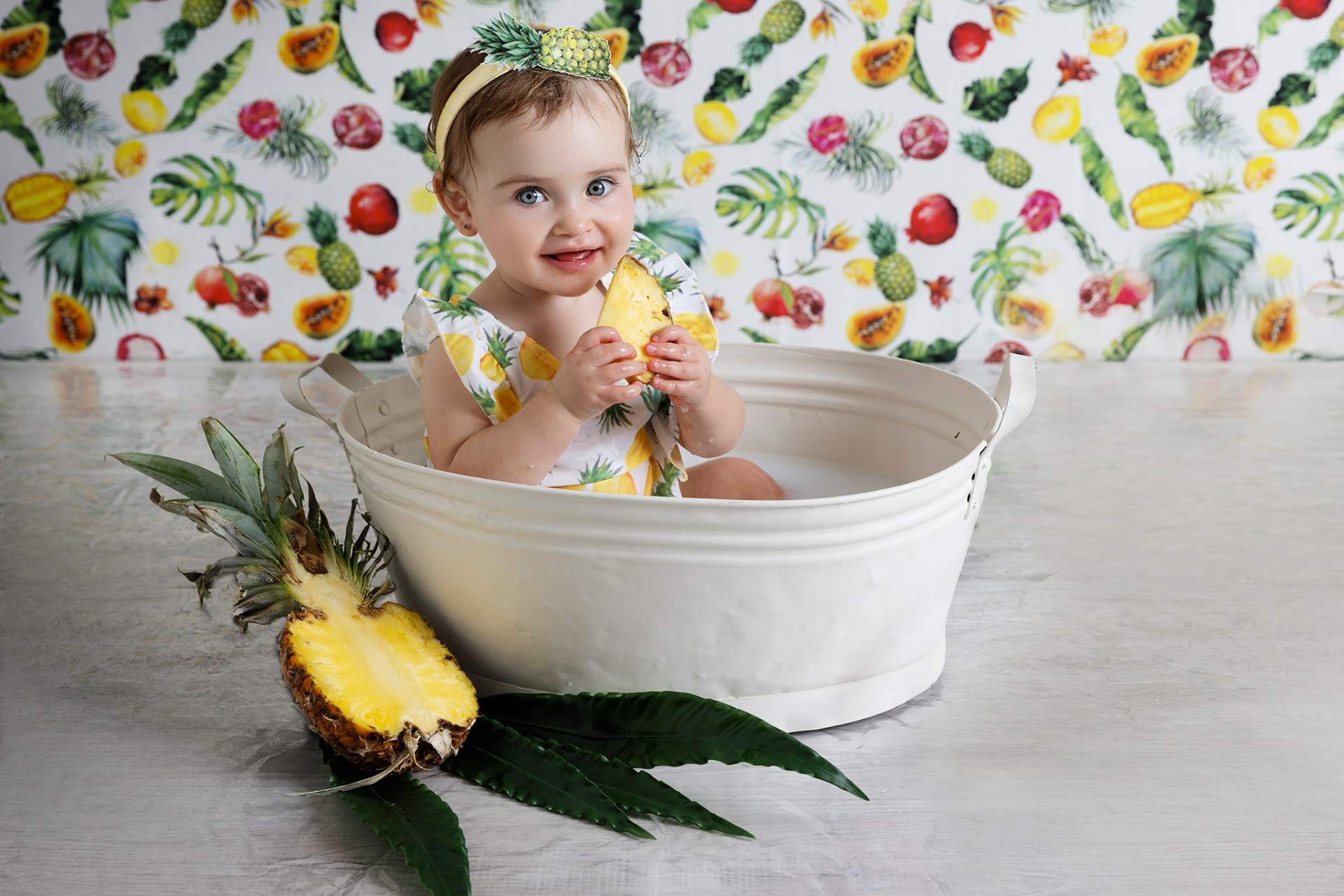 Bambina con outfit a tema ananas, seduta in vasca bianca mentre mangia una fetta di ananas. Sfondo con frutti tropicali e foglie verdi. Foto di primo compleanno realizzata da Claudia Spaziani nel suo studio fotografico a Bologna.