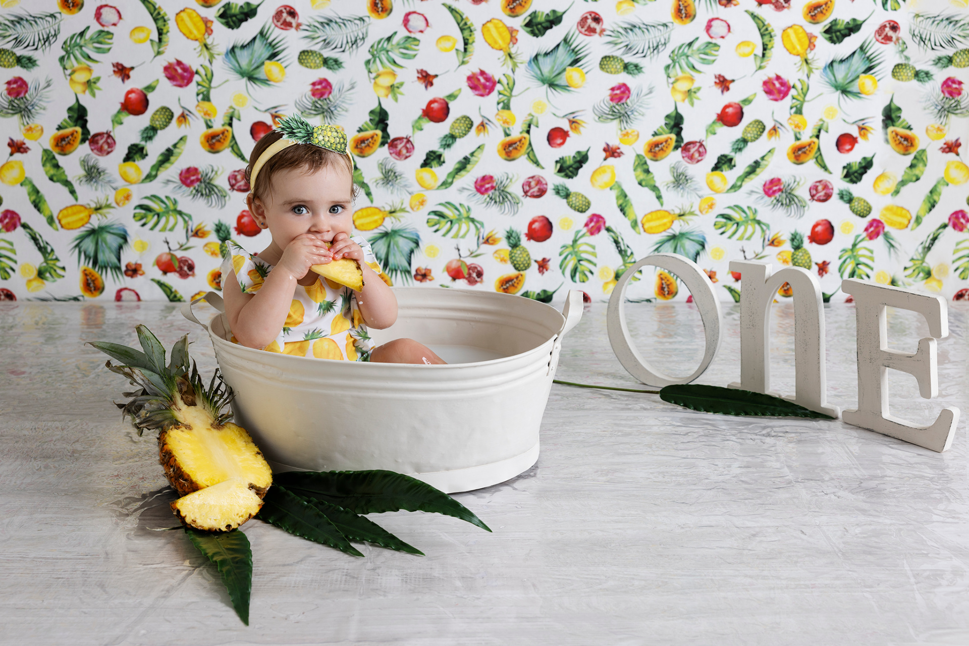 Bambina con vestito giallo e fascia ananas, seduta in vasca bianca con fetta di ananas. Sfondo fruttato e lettere "ONE" indicano il primo compleanno. Ritratto creativo scattato da Claudia Spaziani nel suo studio fotografico a Bologna.