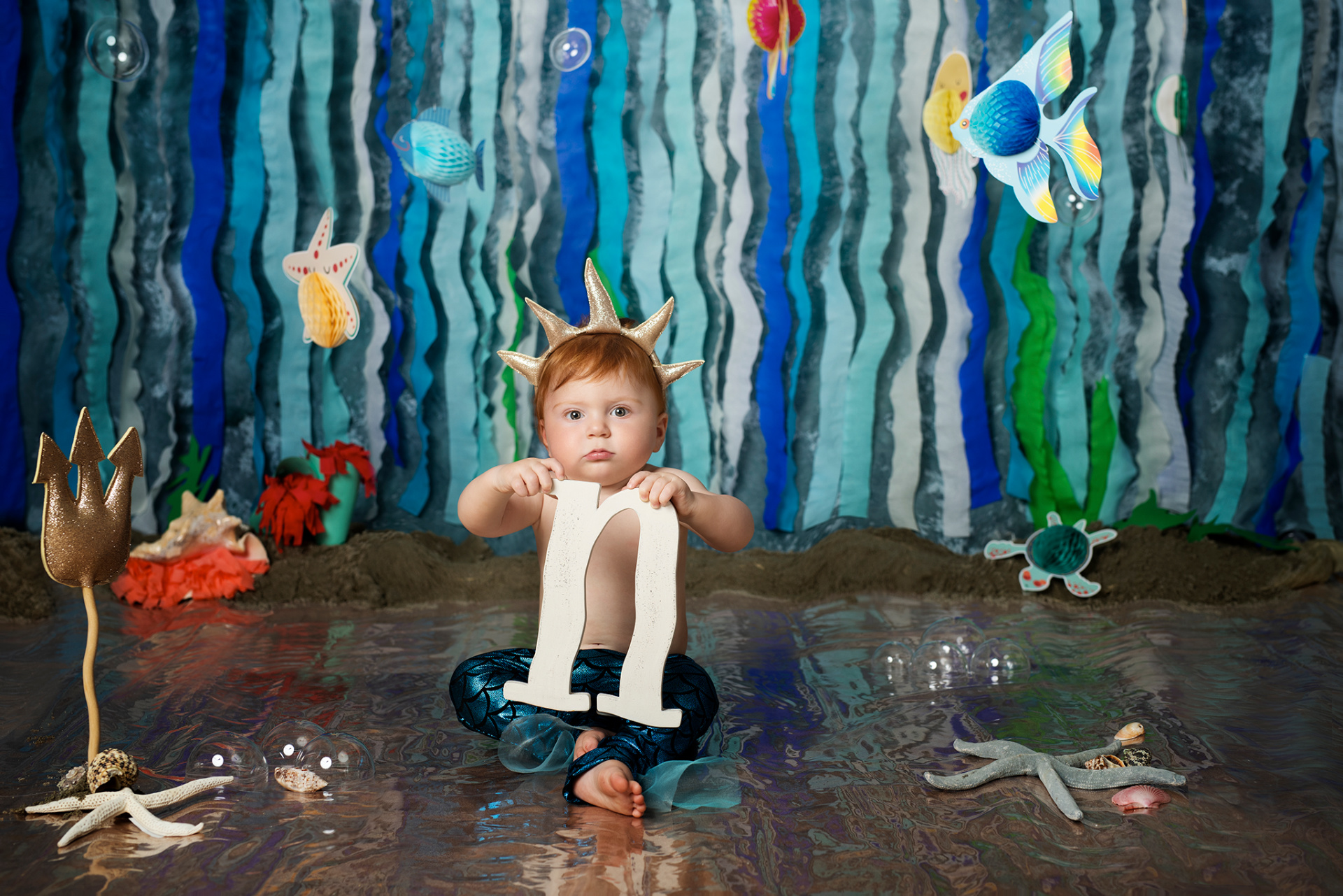 Bambino con corona dorata e pantaloni a squame blu, tiene la lettera "n" bianca. Sfondo marino con creature acquatiche e tridente dorato. Foto di primo compleanno a tema Sirenetta realizzata da Claudia Spaziani nel suo studio fotografico a Bologna.