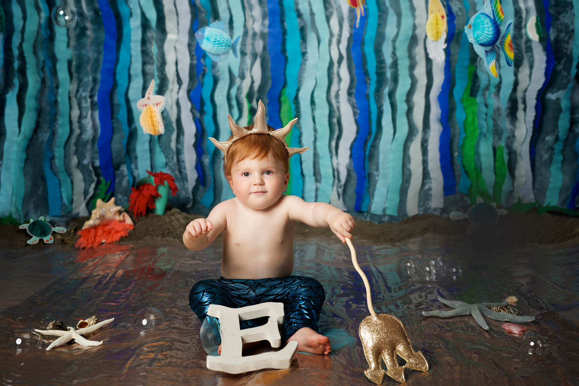 Bambino con pantaloni blu a squame e corona corallo, seduto accanto alla lettera "E" e decorazioni marine. Ritratto di primo compleanno ispirato alla Sirenetta, scattato da Claudia Spaziani nel suo studio fotografico a Bologna.