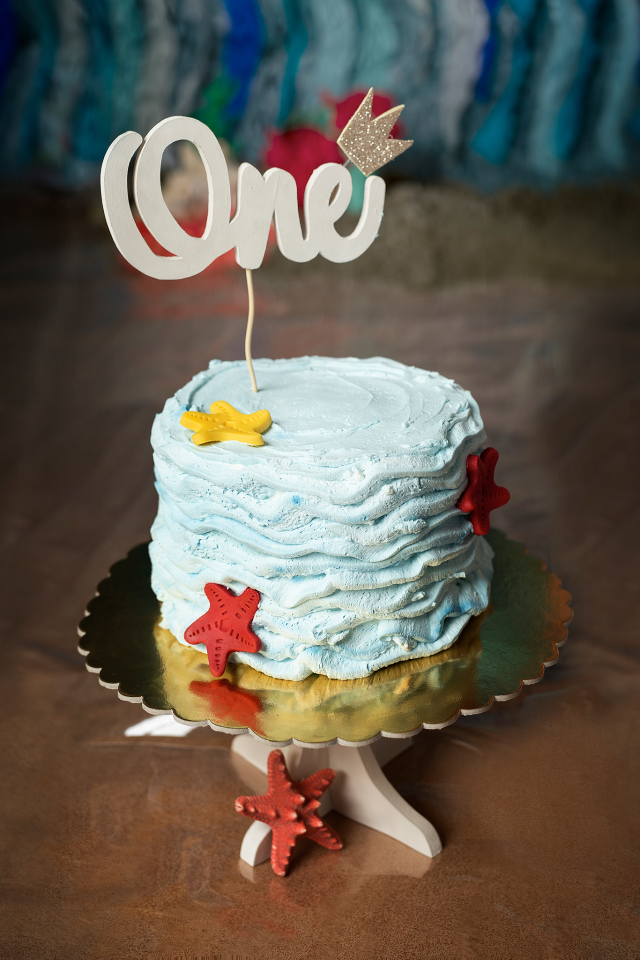 Torta azzurra con decorazioni a onde e stelle marine in pasta di zucchero, topper "One" con corona glitterata. Foto di torta di primo compleanno a tema Sirenetta, realizzata da Claudia Spaziani nel suo studio fotografico a Bologna.