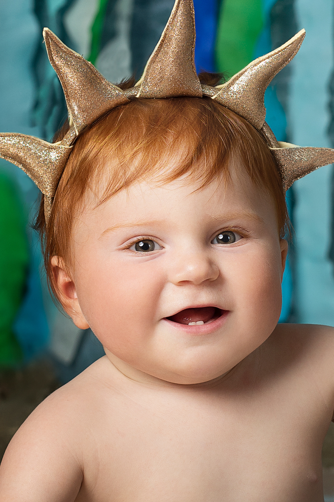 Bambino con capelli rossi e corona dorata, sorride davanti a sfondo marino con nastri blu e verdi. Ritratto festivo ispirato alla Sirenetta, scattato da Claudia Spaziani nel suo studio fotografico a Bologna.
