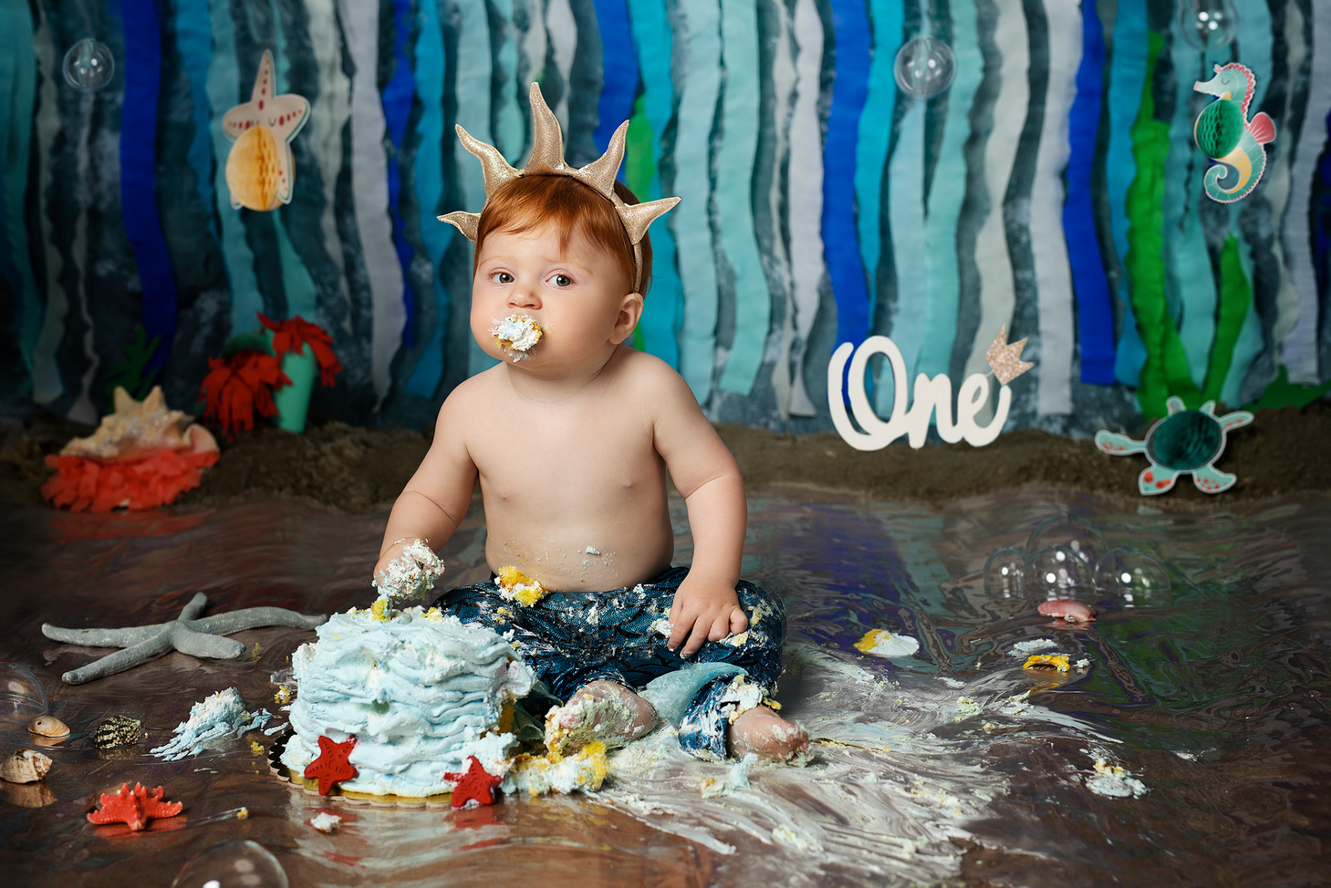 Bambino con pantaloni blu e corona dorata, seduto su pavimento riflettente con torta azzurra distrutta. Sfondo marino con decorazioni subacquee e scritta "One". Ritratto di smash cake a tema Sirenetta realizzato da Claudia Spaziani nel suo studio fotografico a Bologna.