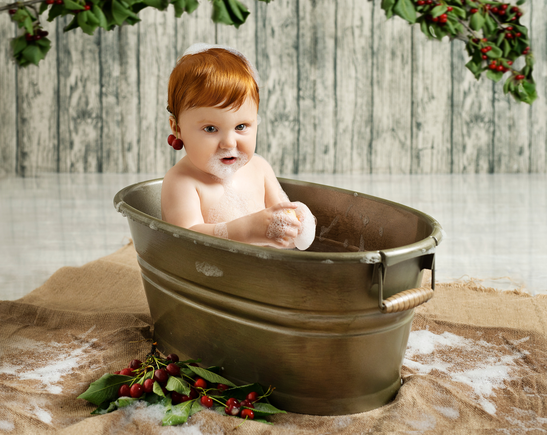 Bambino con capelli rossi seduto in tinozza di metallo piena di schiuma, con spugna in mano e bolle di sapone. Sfondo rustico in legno e decorazioni natalizie con bacche rosse. Ritratto creativo di bagno festivo realizzato da Claudia Spaziani nel suo studio fotografico a Bologna.
