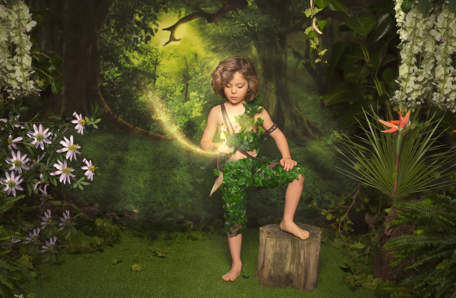 Bambino con costume di foglie verdi e piede su ceppo, circondato da fiori e luce magica. Interazione con figura luminosa volante. Foto narrativa ispirata a Peter Pan scattata da Claudia Spaziani a Bologna.