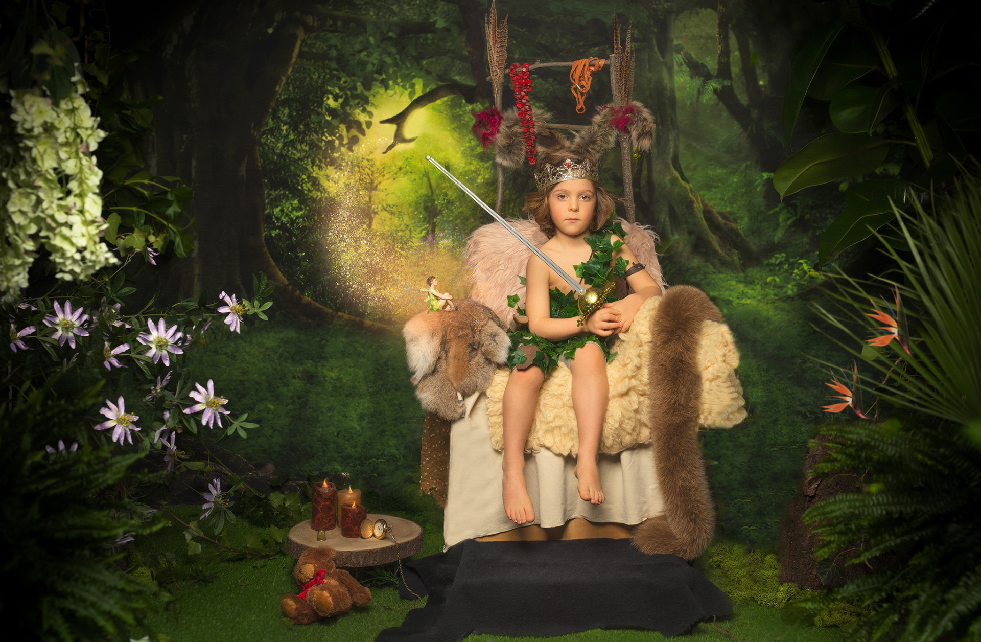 Bambino con corona piumata e spada luminosa, seduto su trono nella foresta con fata sul bracciolo. Oggetti magici come orologio, candele e orsacchiotto completano la scena. Ritratto fantasy ispirato a Peter Pan, realizzato da Claudia Spaziani nel suo studio fotografico a Bologna.
