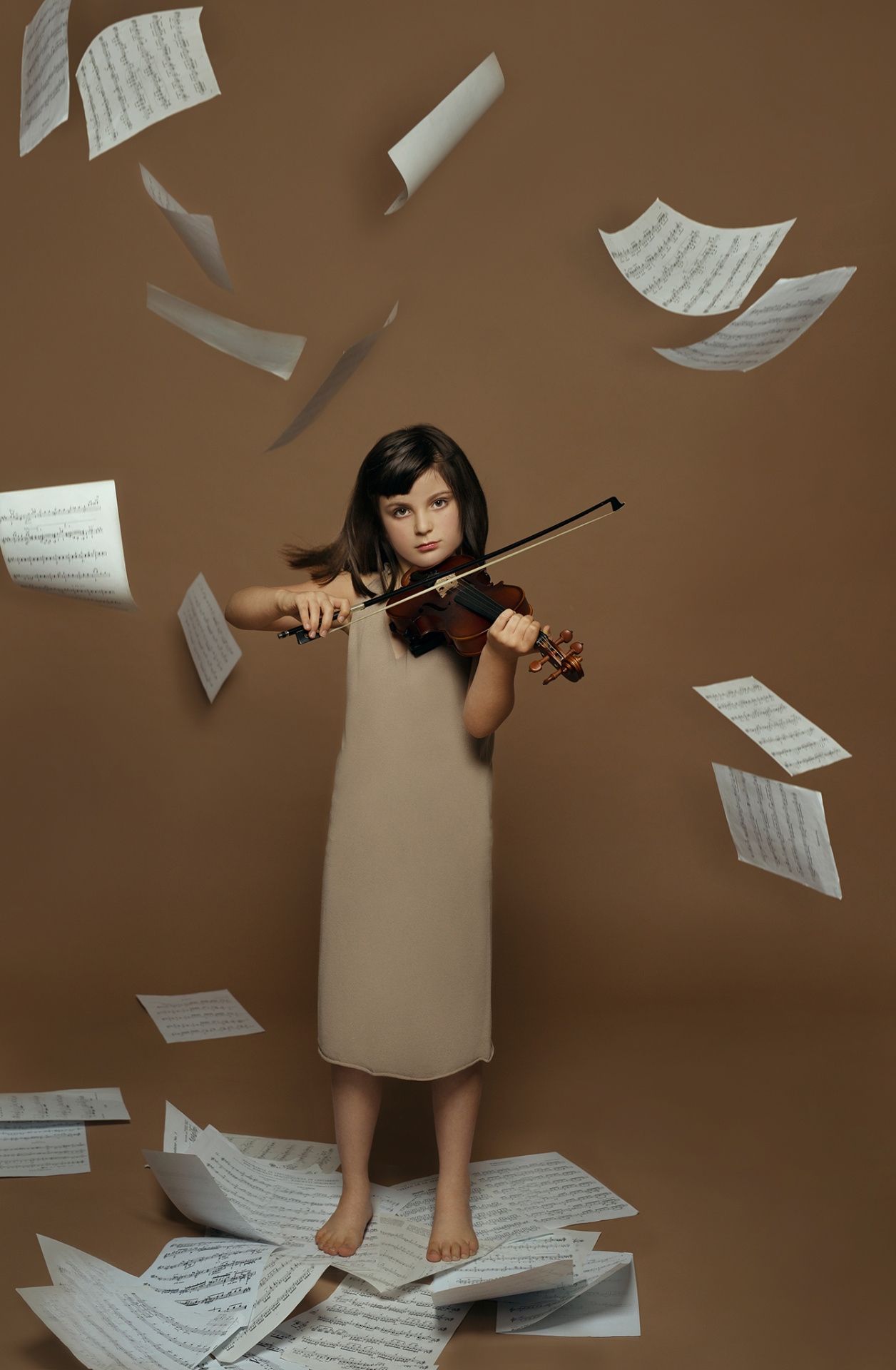 Bambina a piedi nudi su spartiti sparsi, suona il violino circondata da fogli musicali sospesi. Sfondo marrone e atmosfera dinamica. Ritratto espressivo a tema musica e creatività, scattato da Claudia Spaziani nel suo studio fotografico a Bologna.