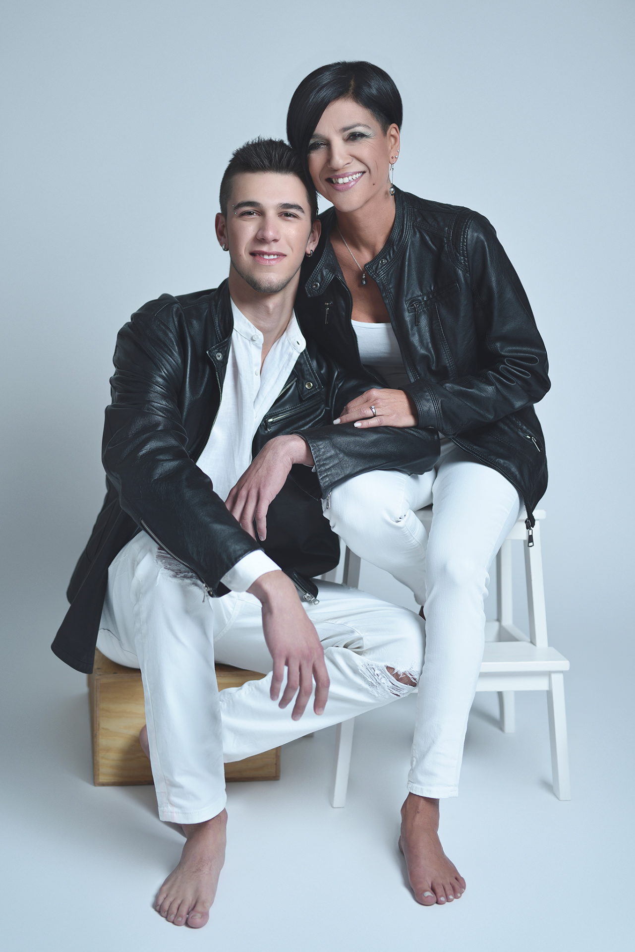 Madre e figlio seduti in studio indossano giacche di pelle nere e abiti bianchi coordinati, in un ritratto fine art realizzato da Claudia Spaziani nel suo studio fotografico a Bologna.