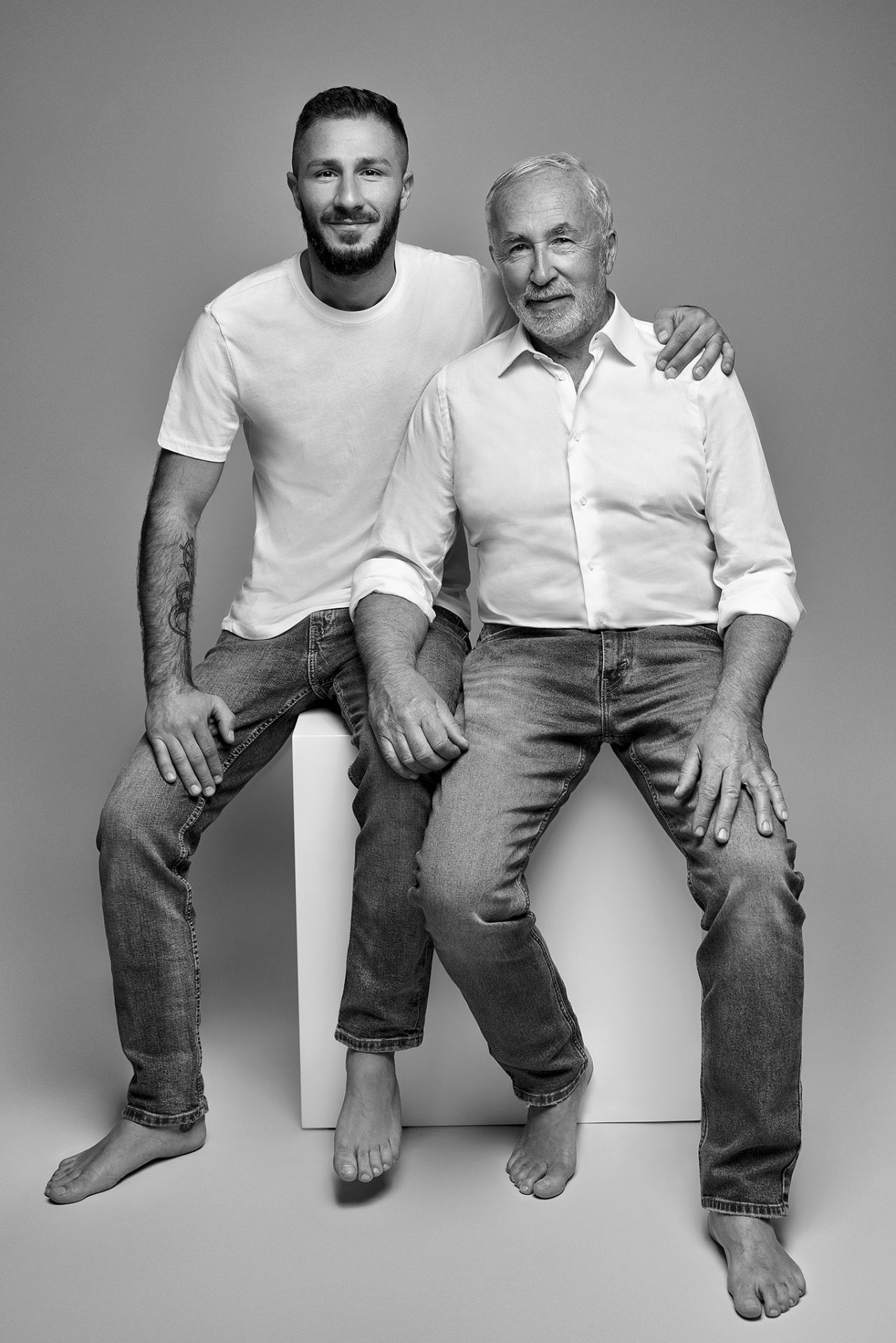 Fotografia fine art in bianco e nero di padre e figlio seduti su cubo bianco, scattata da Claudia Spaziani a Bologna. Entrambi in jeans e camicie bianche, a piedi nudi, in posa affettuosa e rilassata.