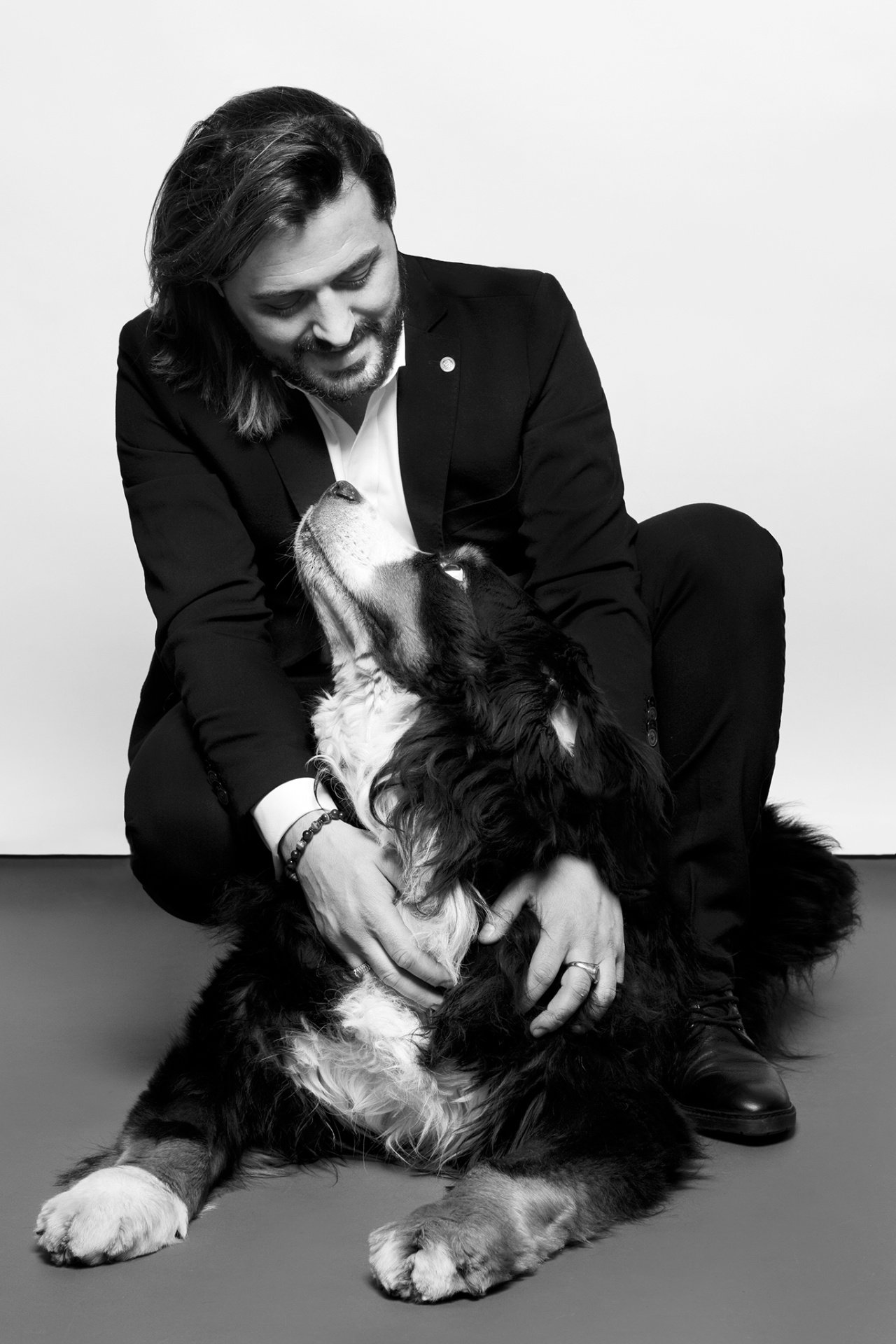 Ritratto in bianco e nero di uomo in abito elegante inginocchiato accanto a Bovaro del Bernese, in un momento di affetto. Fotografia fine art di Claudia Spaziani, Bologna.